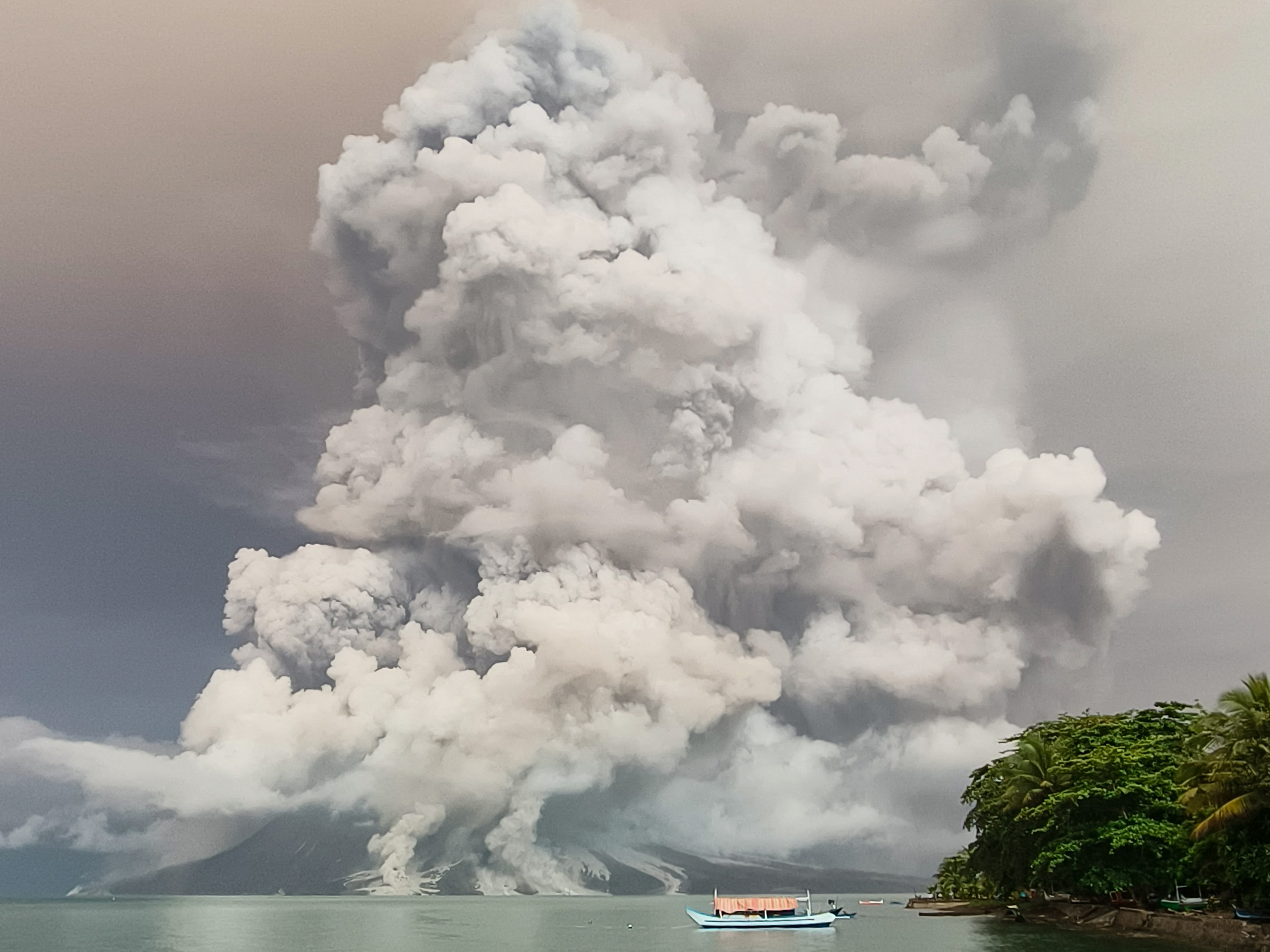 Huge clouds of smoke and ash billowing from the top of the Mount Ruang volcano. The sea is in front, with a boat