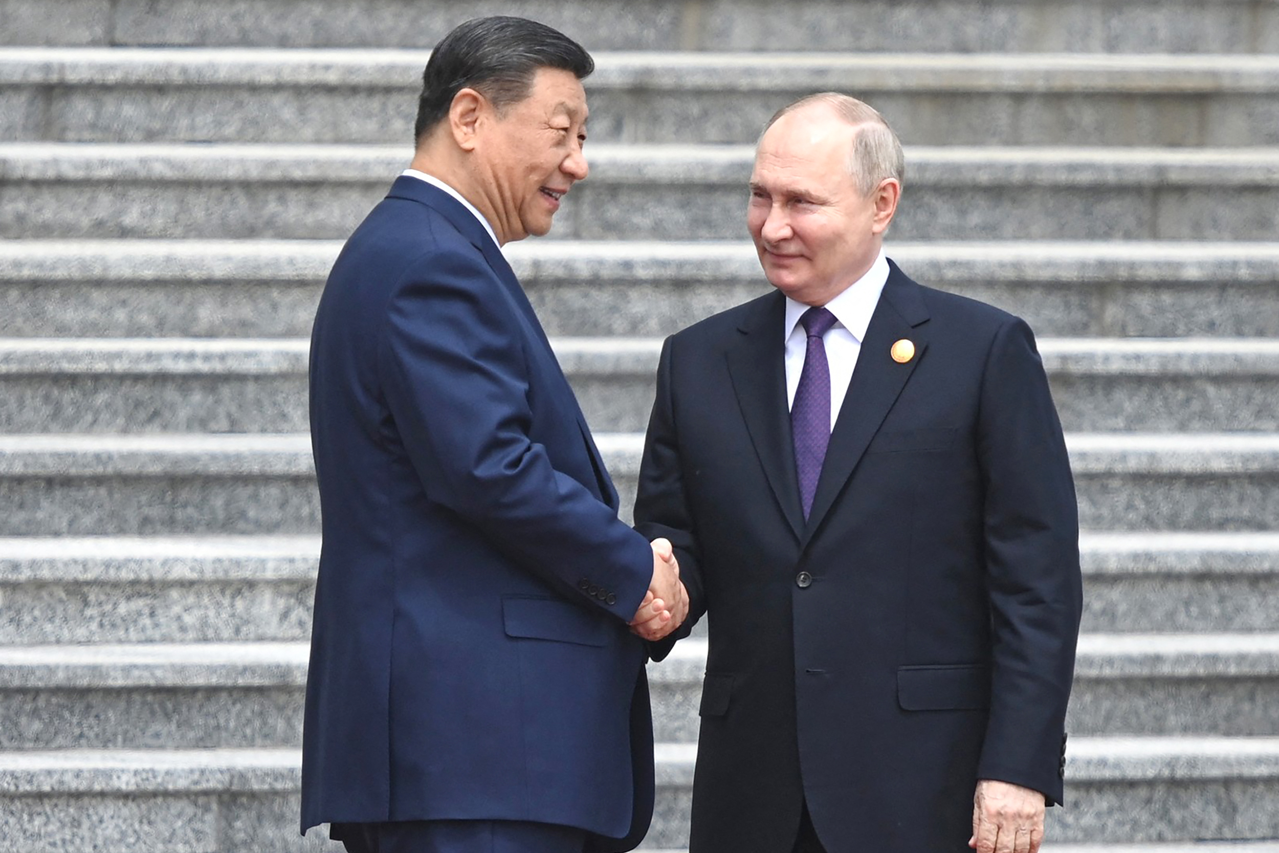 Putin and Xi shaking hands outside the Great Hall of the People. The steps are behind them. They are both smiling.