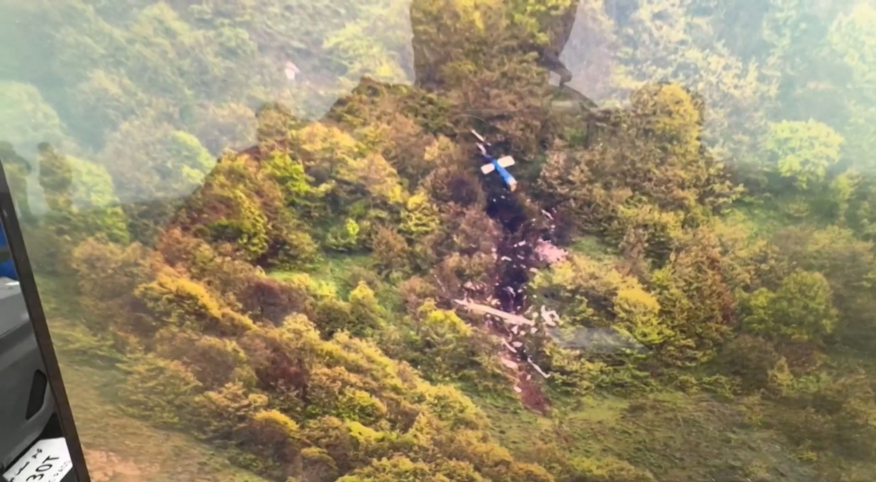 A black burned area on a wooded mountainside. The blue and white tail of a helicopter can be seen.