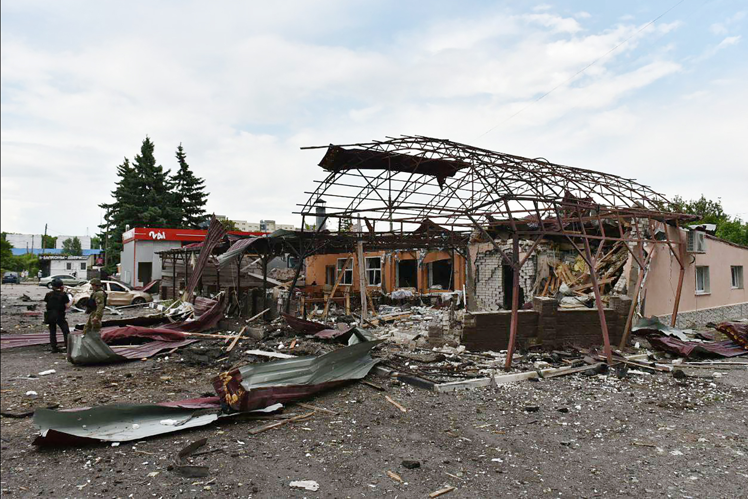 Destroyed buildings in attacks on Kharkiv. There is debris including metal sheets on the ground. There are also damaged cars. Two people in uniform are near the wreckage