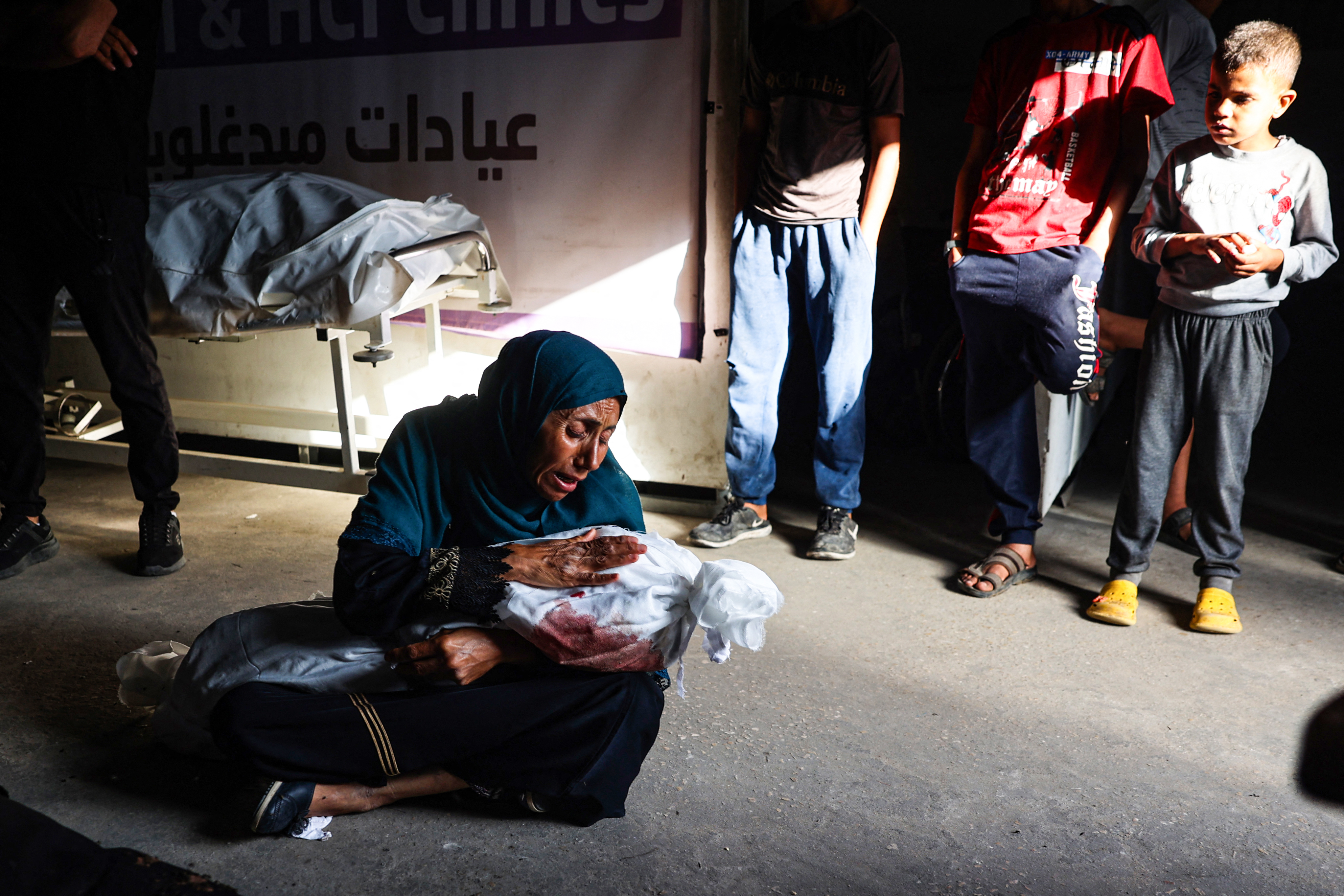 A child looks on as Palestinian woman holds the shrouded body of another child killed in Israeli bombardment