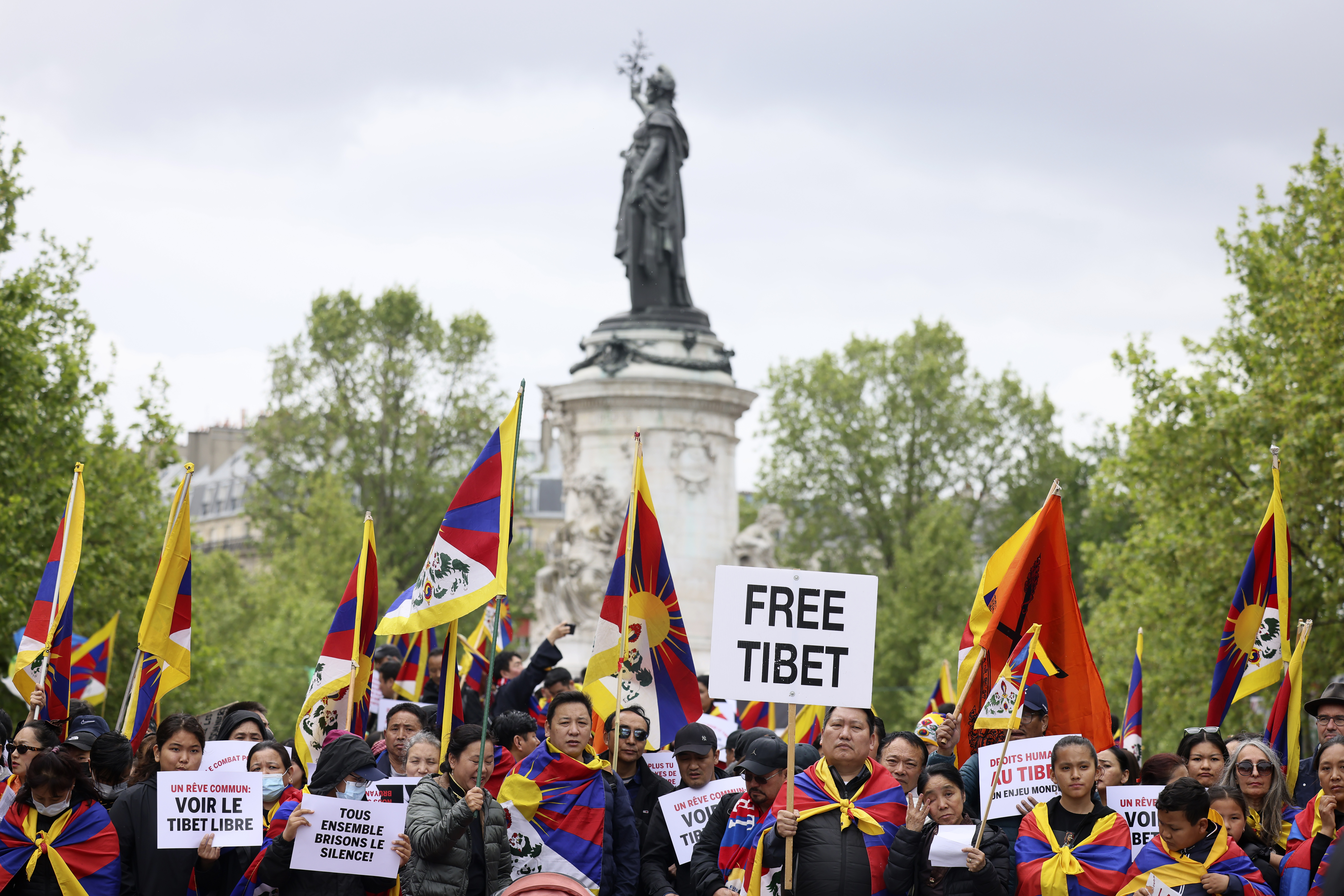 Tibet protesters rally in Paris as Xi Jinping arrives. They have placards reading 'Free Tibet' and are carrying Tibetan flags. There's a statue behind them.