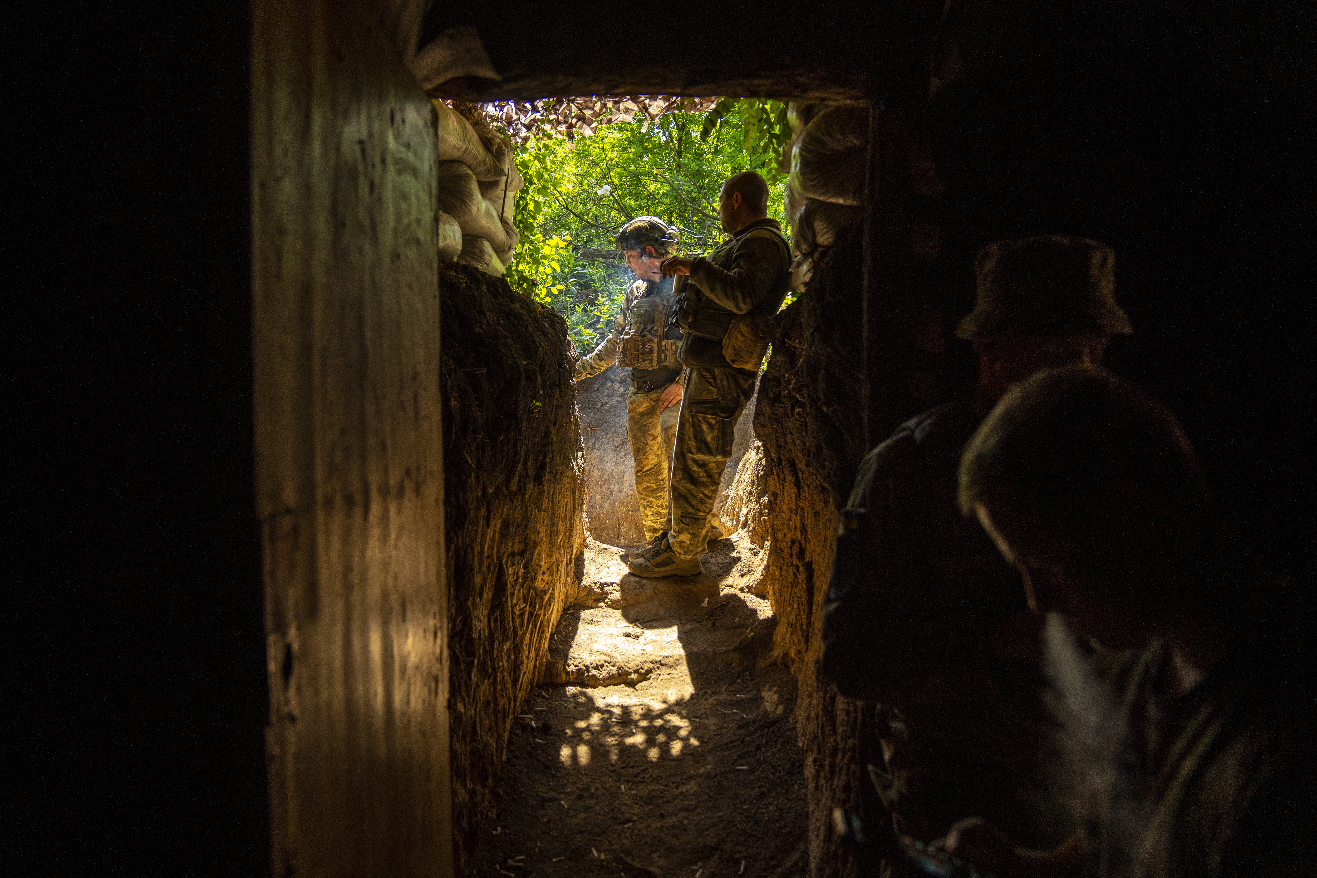 Two Ukrainian soldiers smoke cigarettes outside their trench near Avdiivka. The sun is shining and there is lots of greenery in the background.