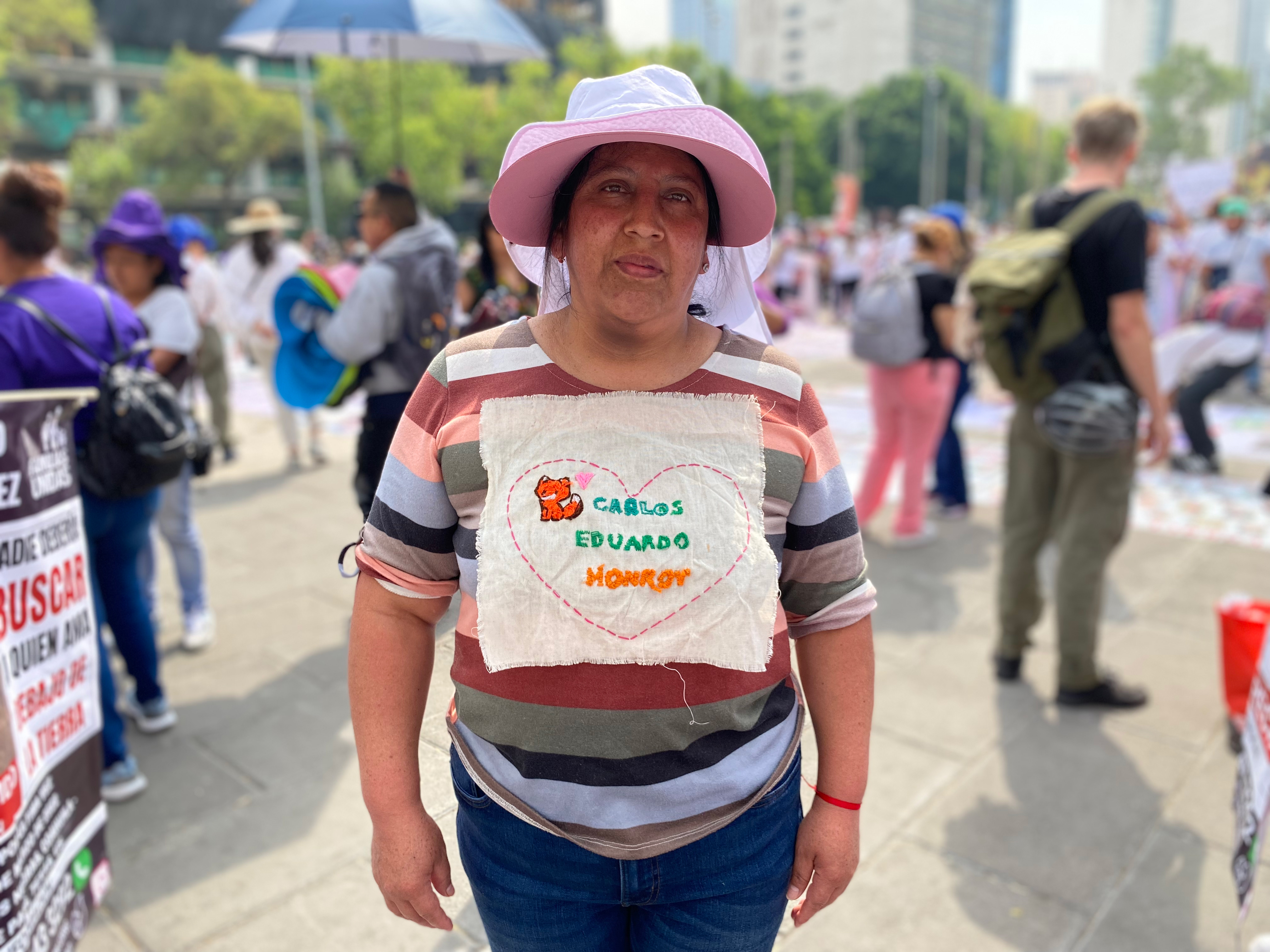 A woman in a pink hat and striped shirt stands with an embroidered message pinned to her chest, containing the name of her missing son surrounded by a heart.