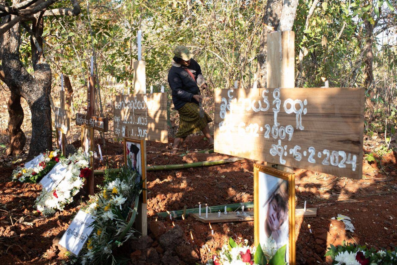 The graves of the four children killed at the Daw Si Ei village school. There is a wooden boafrd in each with their name and wreaths of flowers. Some people are walking away behind