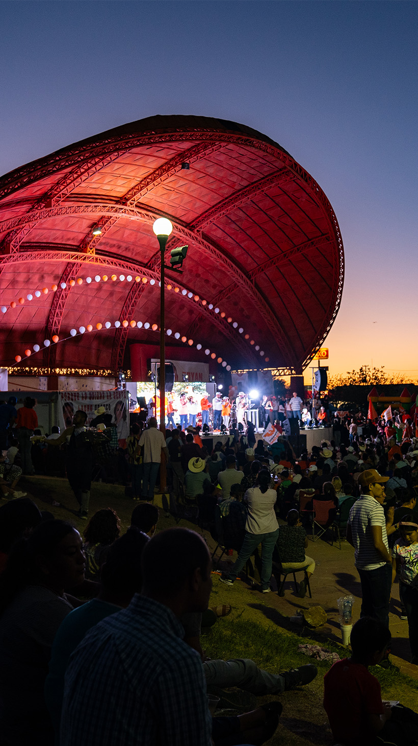 Politicians speak under a bandshell in Mexico at night, a crowd gathering before the stage to hear them.