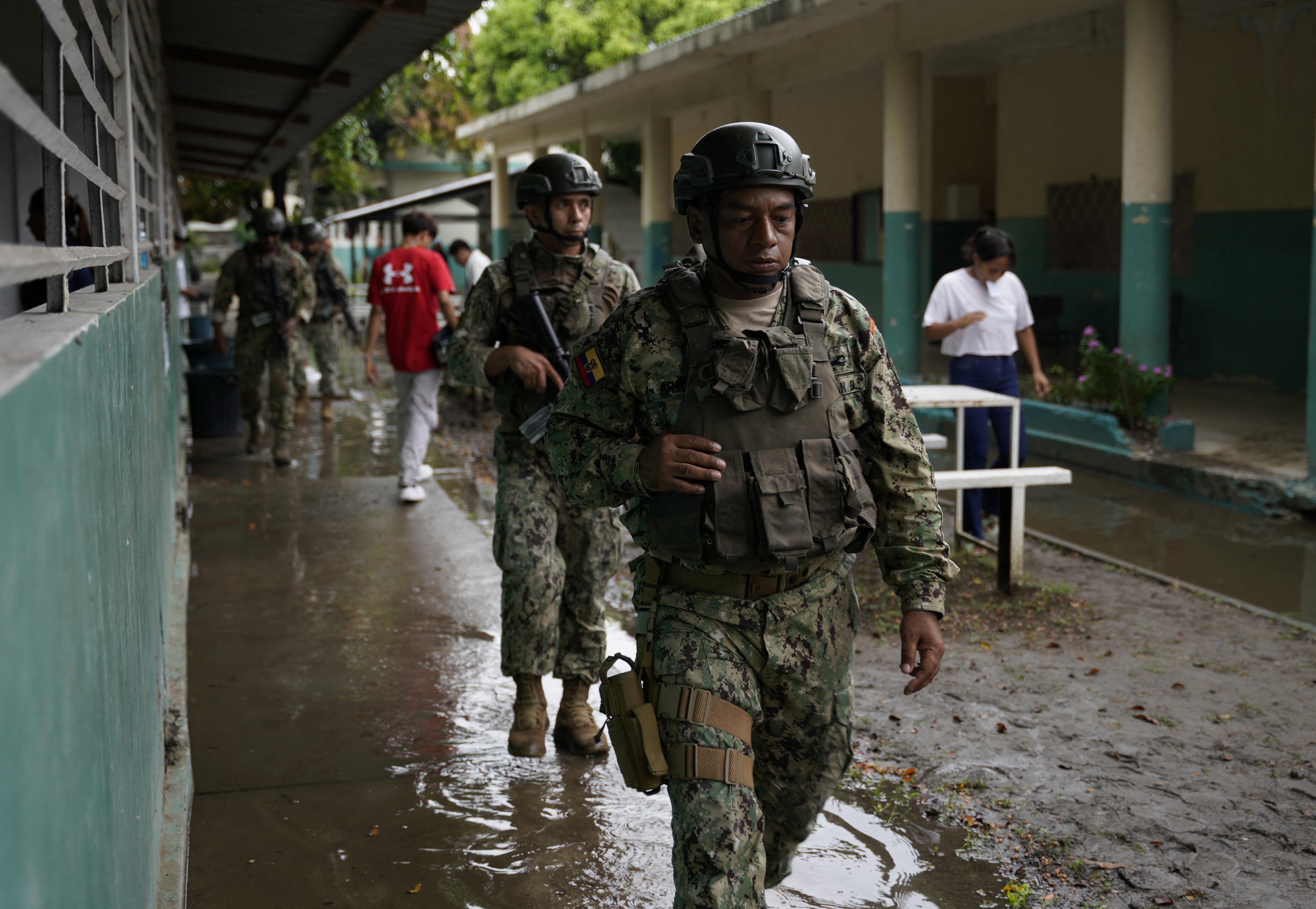 Two soldiers in helmets and fatigues walk in a row down a muddy path between buildings in Guayaquil.