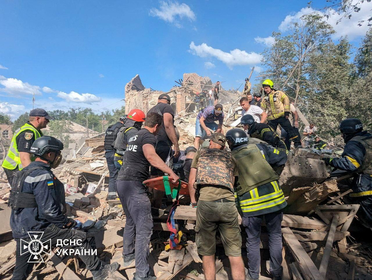 Rescuers at an apartment block in Kryvyi Rih that was hit in a Russina missile attack. Thay are carrying a stretcher and are on a pile of rubble and timber. What's left of the building is behind them.