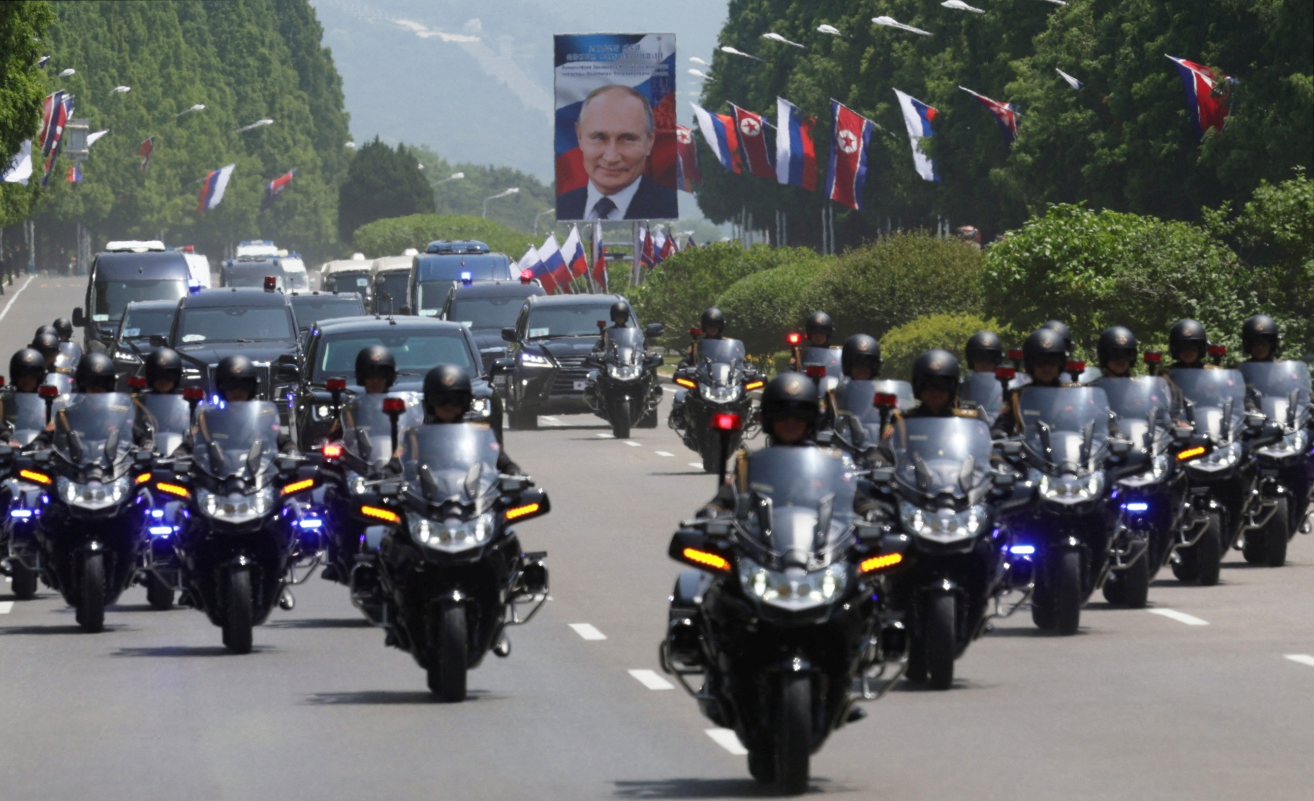 Motorcycle outriders in a V formation lead Putin's limousine through Pyongyang. There are Russian flags and large portraits of Putin along the side of the road