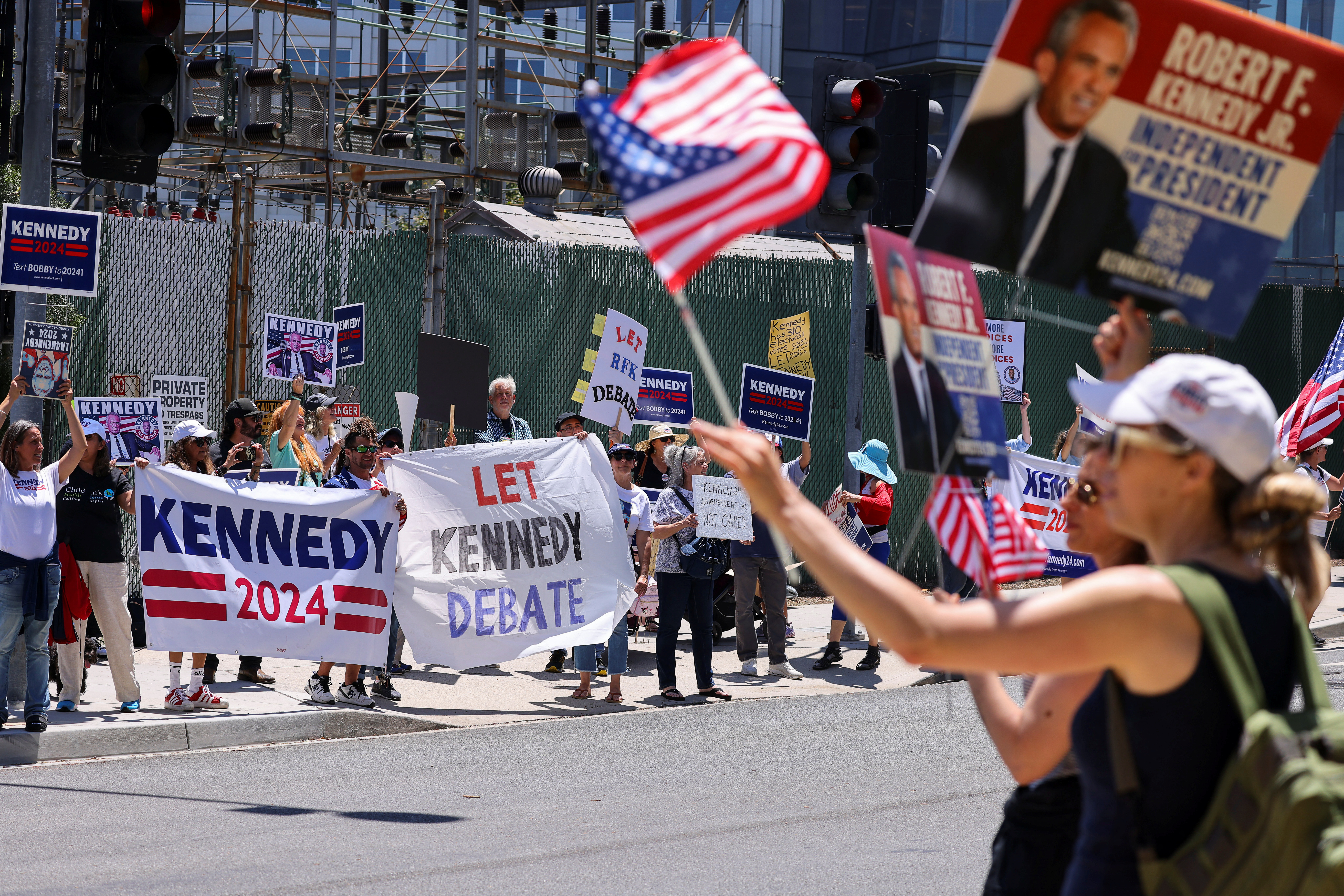 A woman waves an American flag at a rally for Robert F Kennedy Jr, where another person holds up a banner that reads: "Let Kennedy Debate"