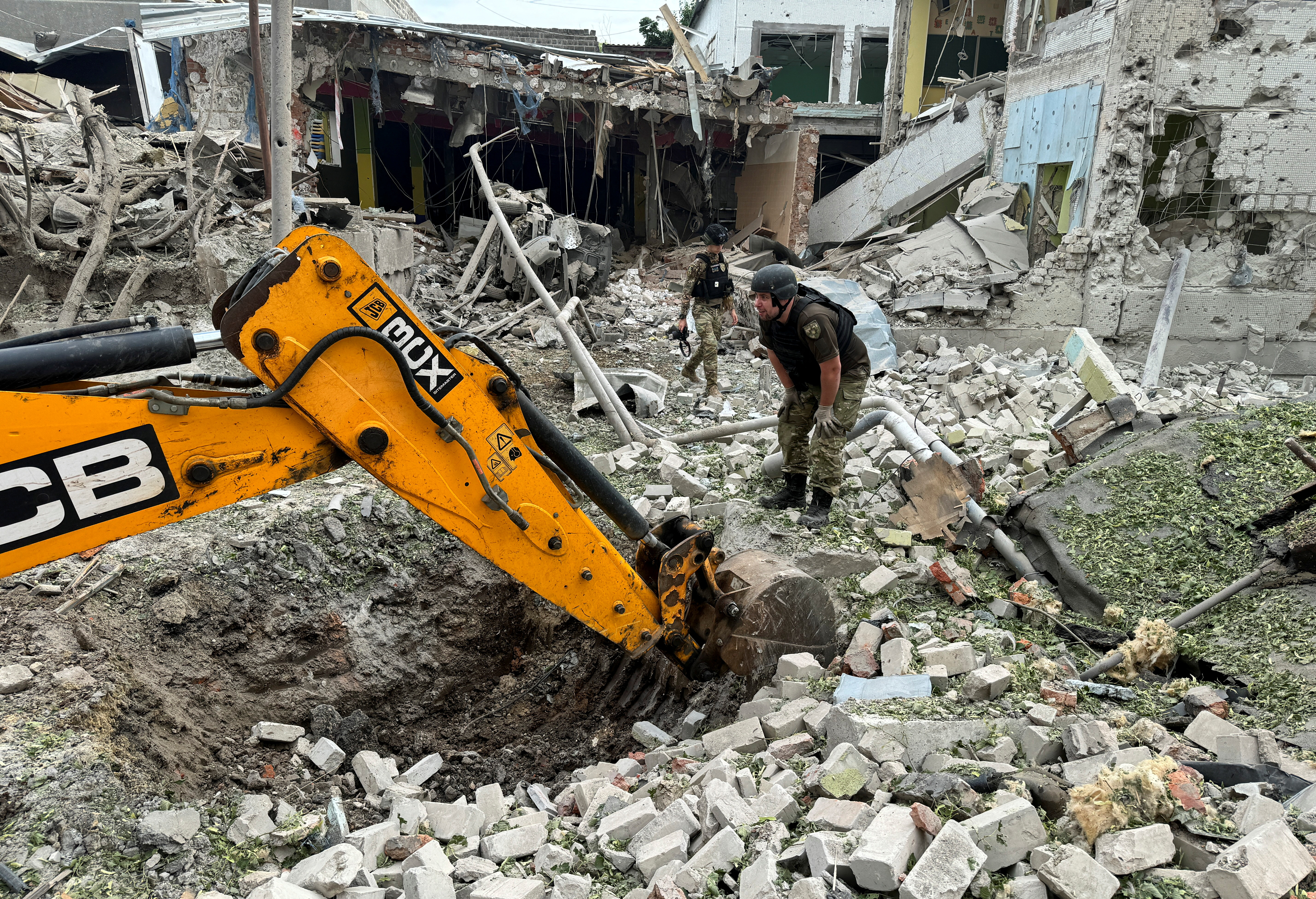 A member of the bomb squad working at the scene of a Russian attack on Kharkiv on Sunday. The officer is standing on a pile of rubble with the destroyed building behind. An excavator is at work.