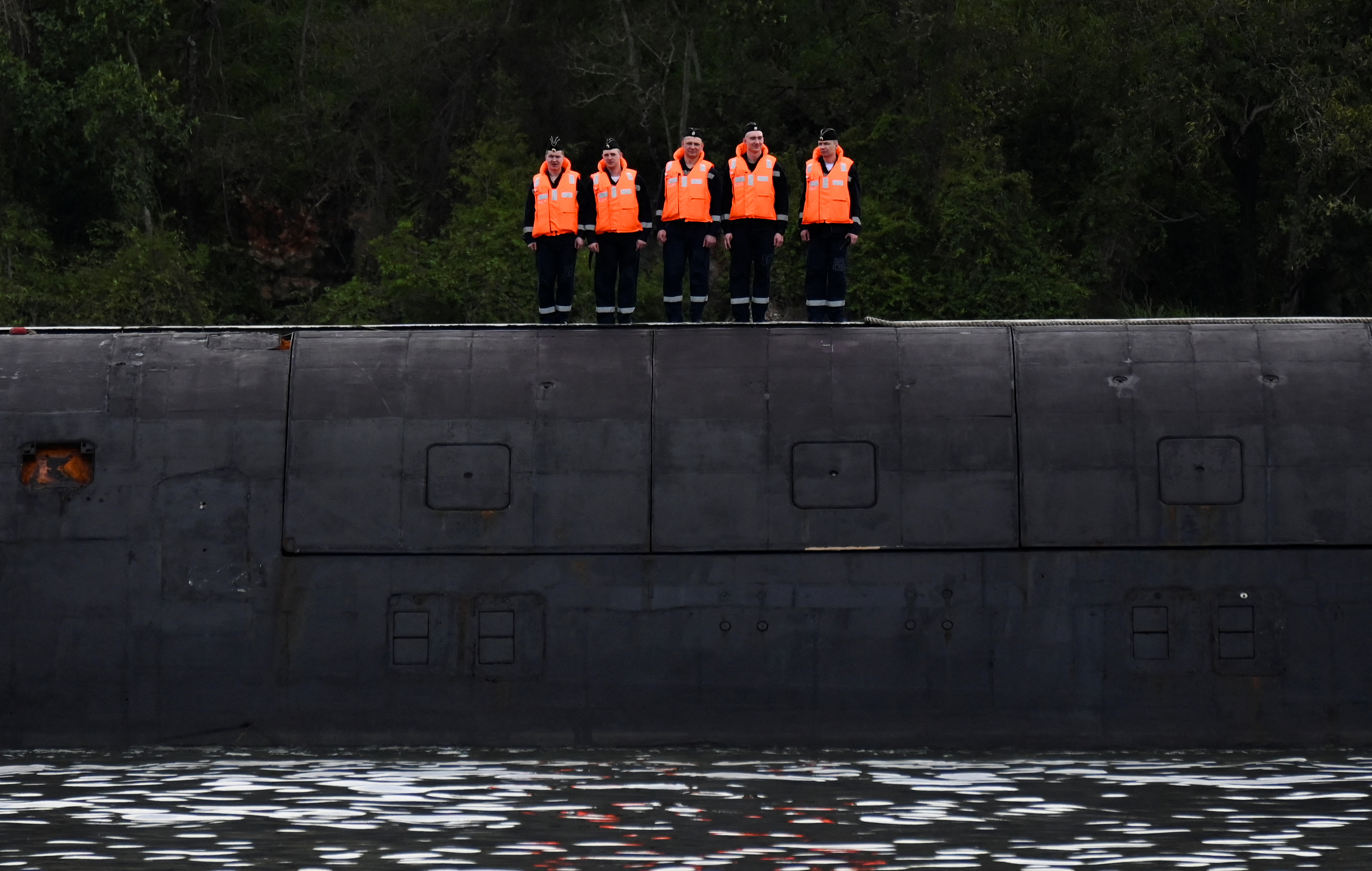 Russian sailors stand guard on top of the nuclear-powered submarine Kazan. They are wearing orange lifejackets, The submarine is black.