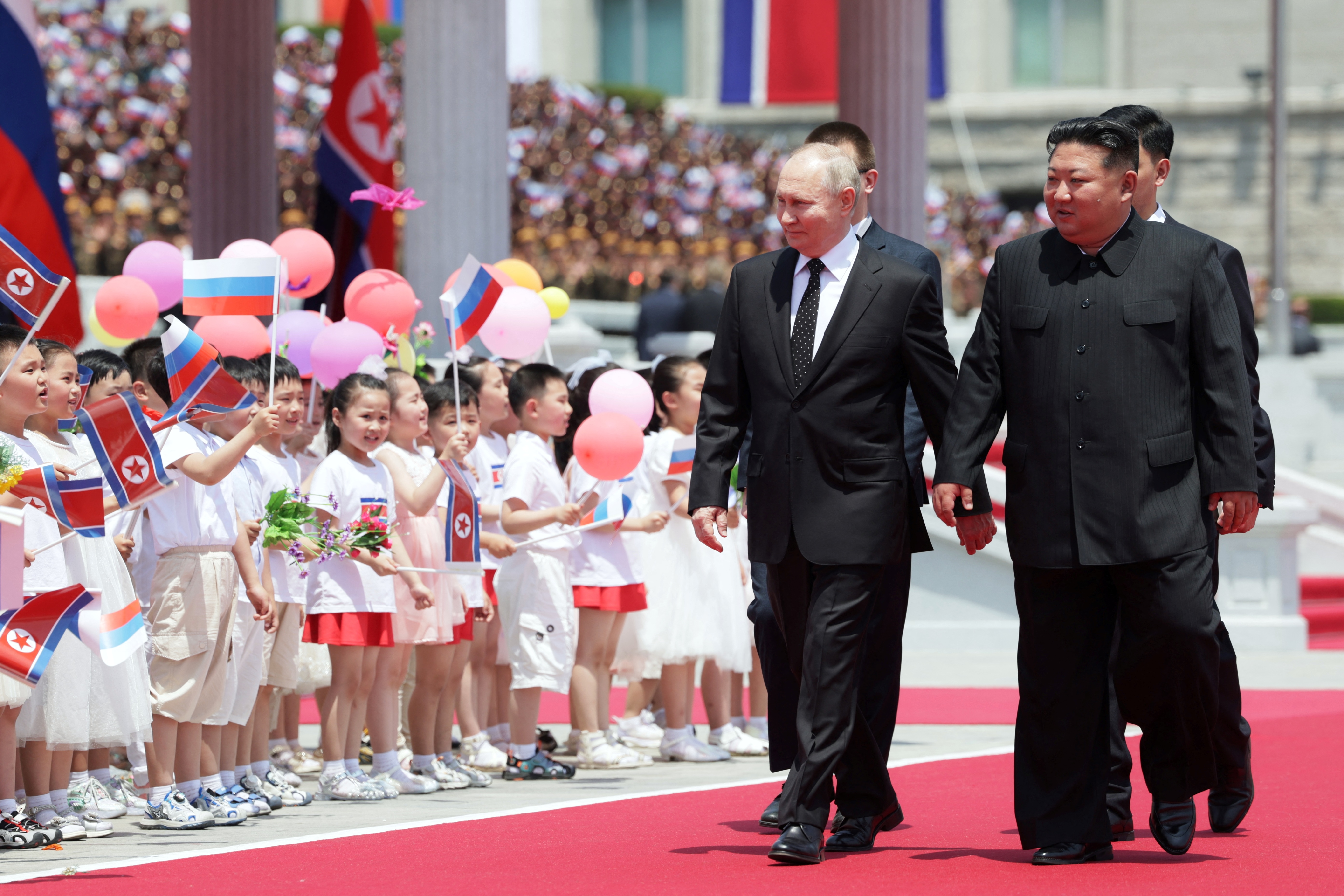 Putin and Kim walk in Kim Il Sung square as young children hold balloons and wave Russian and North Korean flags.