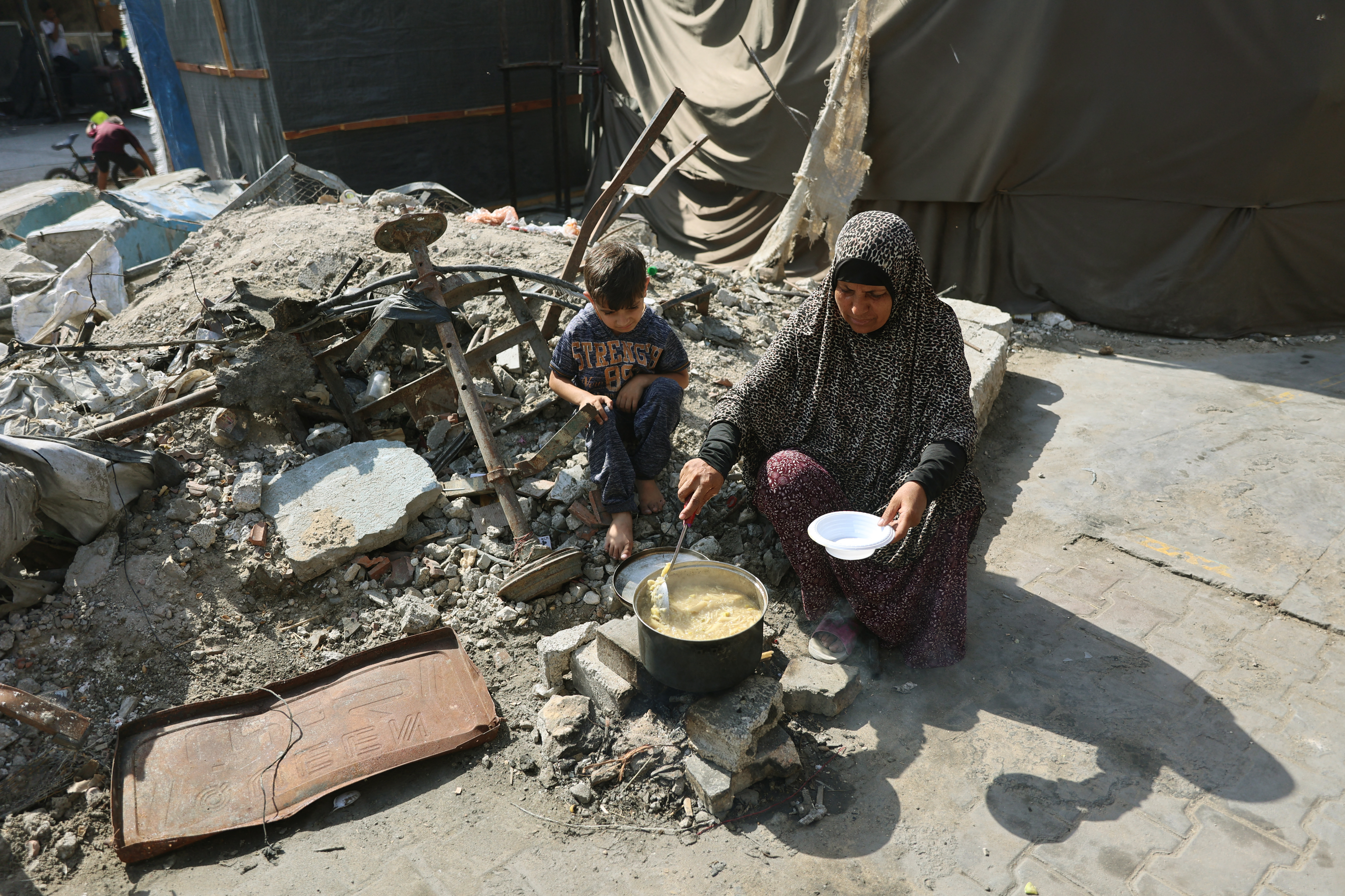 A Palestinian woman sits on some rubble as a child watches her cooking on an open wood fire in the Jabalia refugee camp