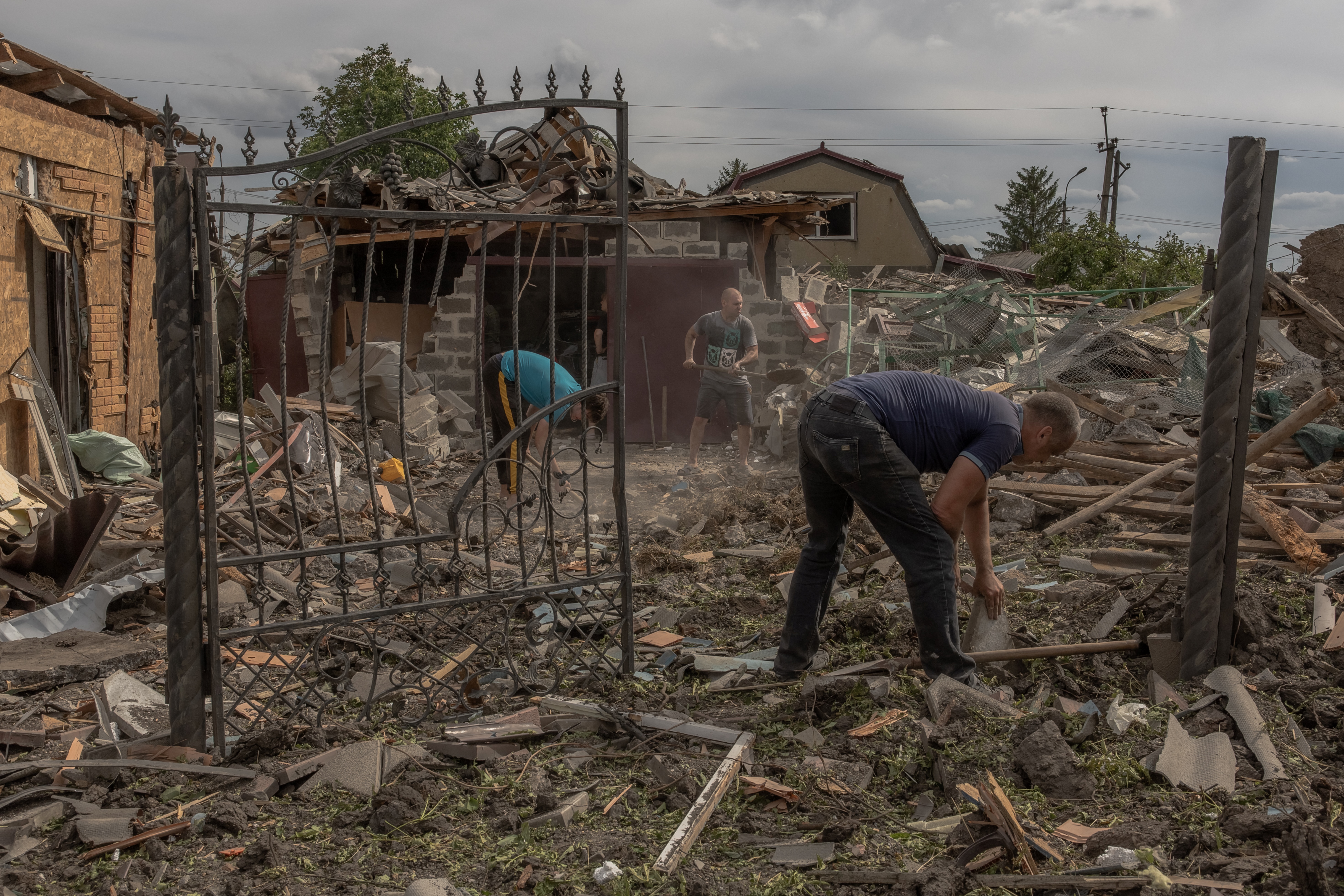 Two men amid rubble from destroyed houses trying to clean up. Half the metal gate is hanging open and is bent. The other side is missing.