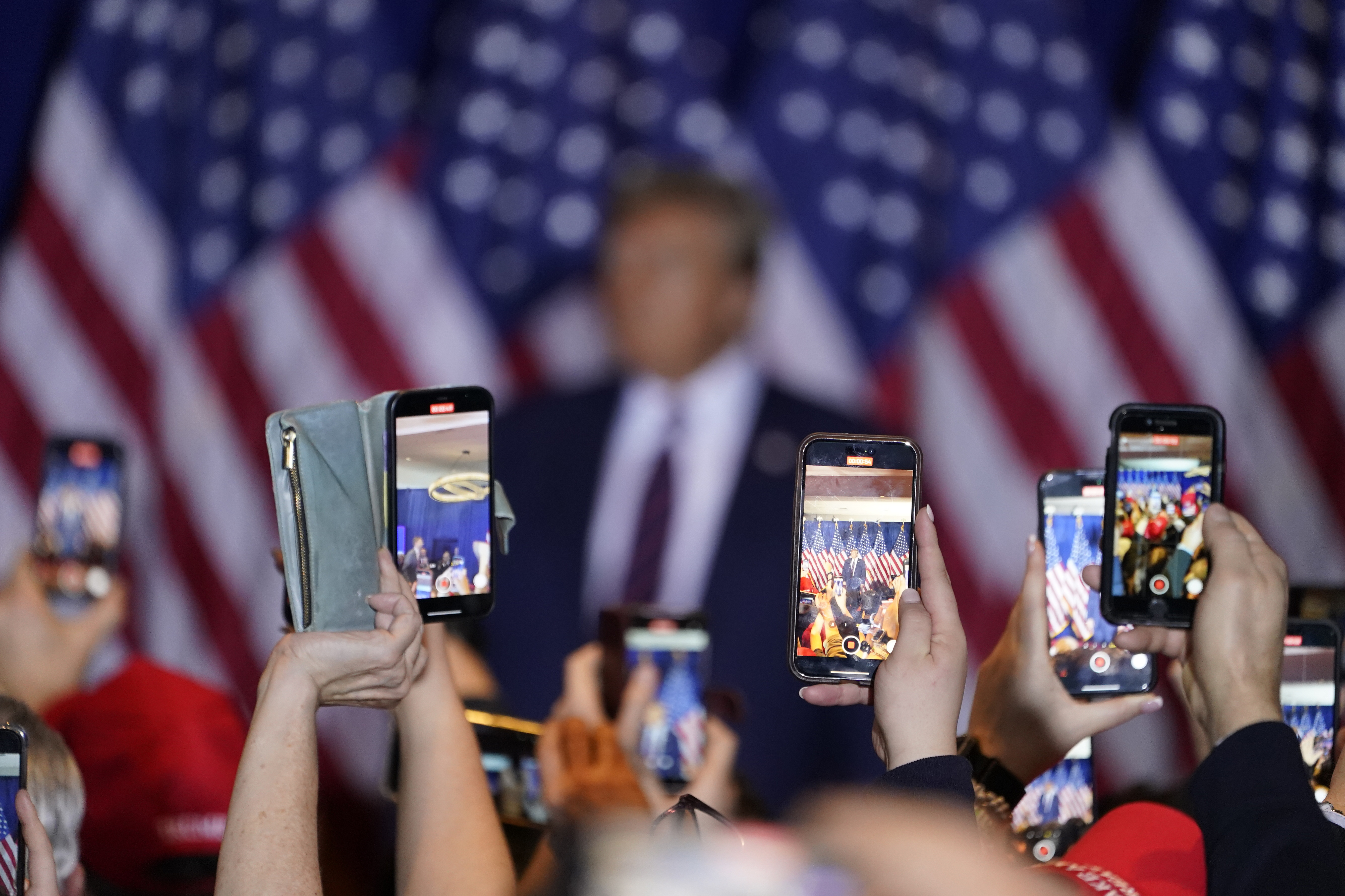 Supporters hold up their cellphones to take pictures of Donald Trump on the campaign stage, surrounded by US flags.