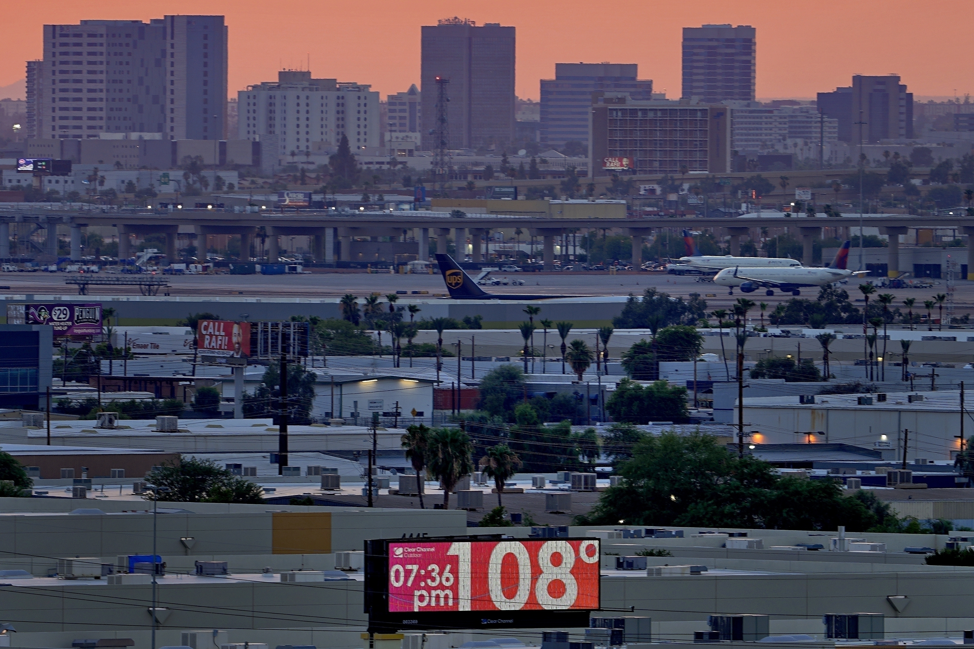 An electronic billboard shows the temperature to be 108 degrees Fahrenheit. Behind the billboard, the skyline of Phoenix, Arizona, is lit by an orange sunset.