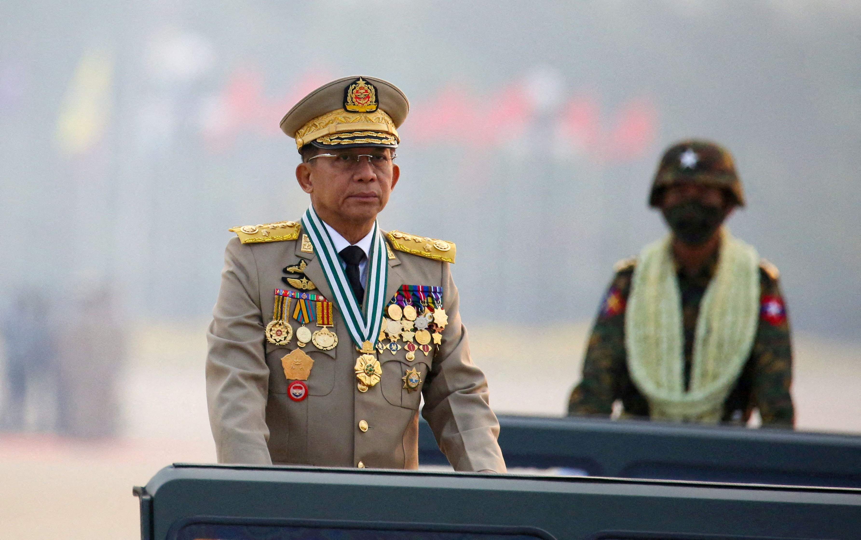 Myanmar Senior General Min Aung Hlaing. He's standing in a open topped jeep. He is wearing a military uniform with lots of medals. Another soldier is behind him.