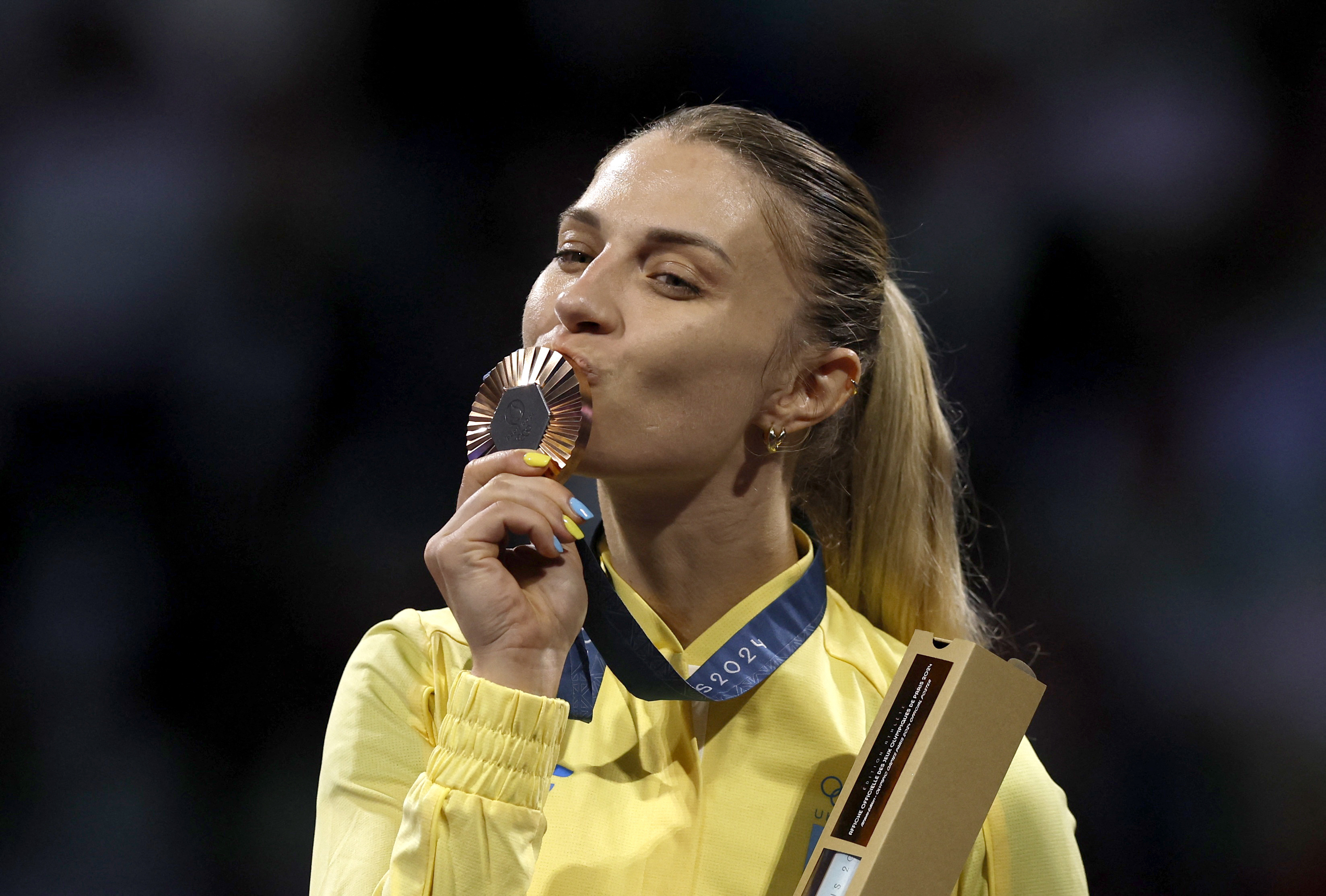 Olga Kharlan kisses her bronze medal. She has long blonde hair in a pony tail and is wearing a yellow jacket. She looks very happy.