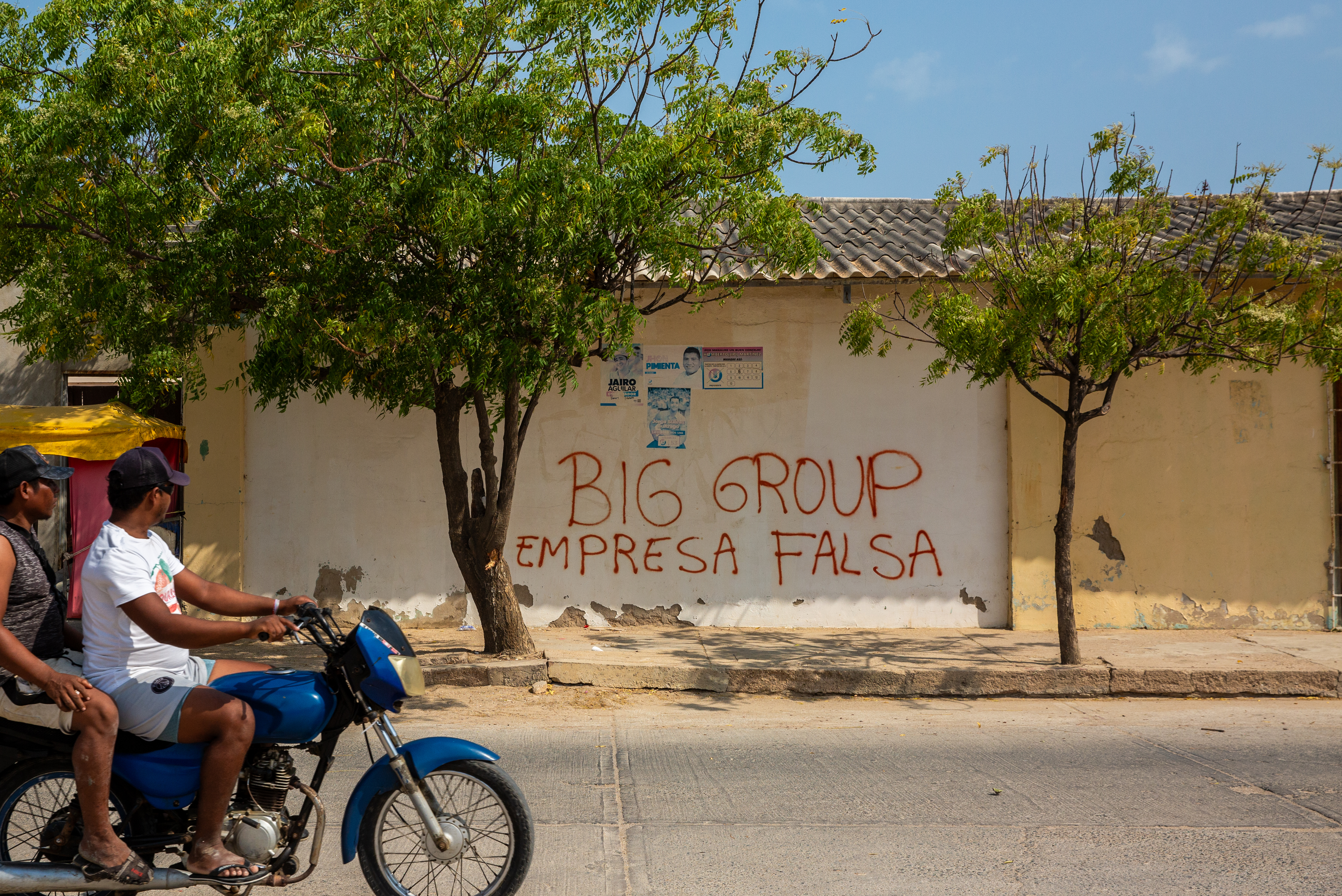 Two men on a motorcycle pass graffiti that reads in Spanish: 