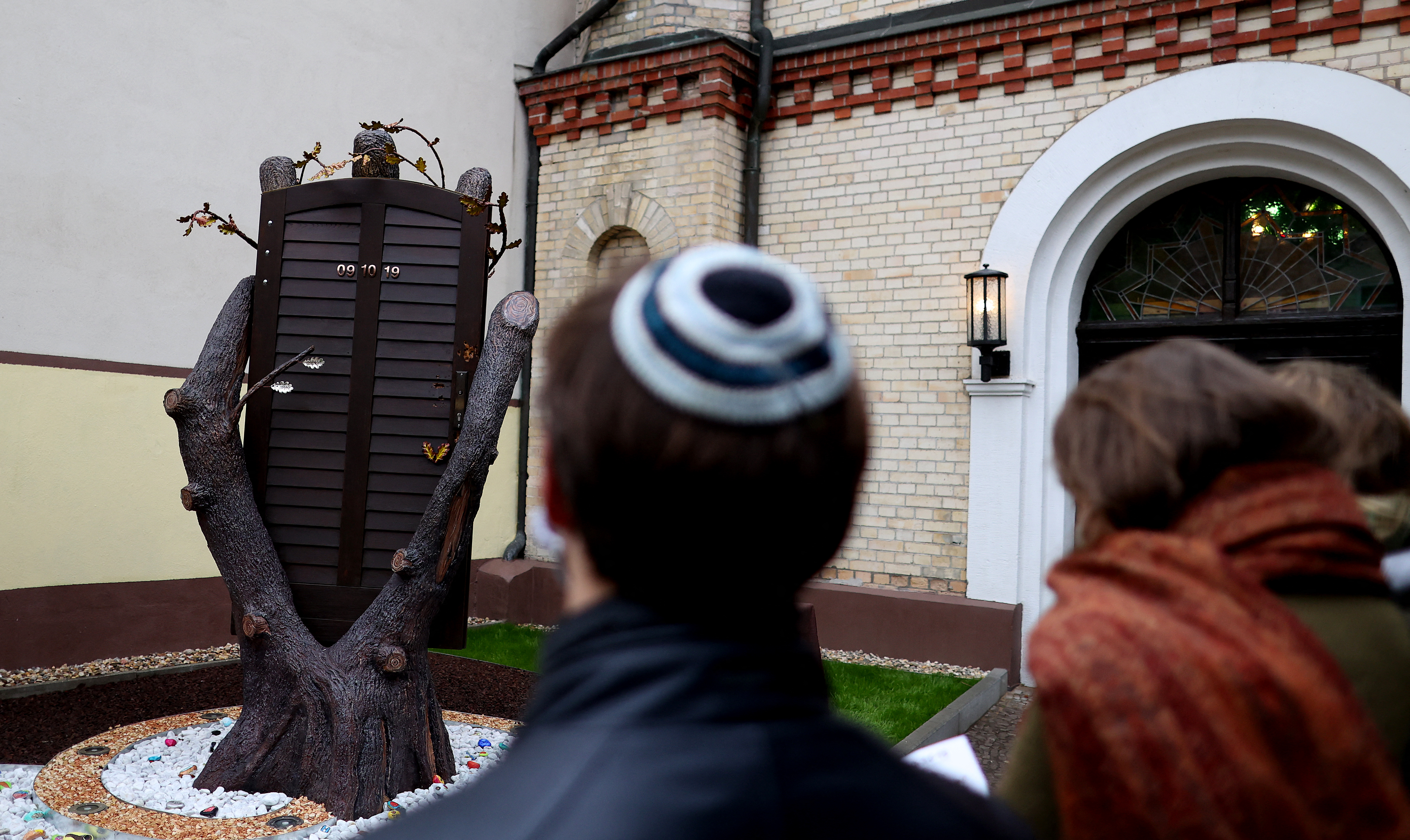 People stand next to a new memorial made of the original entrance door inside the synagogue in Halle, eastern Germany, on October 09, 2020, on the first anniversary of the anti-Semitic attack on the synagogue. Last year's attack on the synagogue in Halle came on October 9 during Yom Kippur, the holiest day in the Jewish calendar, and was one of the worst acts of anti-Semitic violence in Germany's post-war history. Two people were shot dead after an extremist tried and failed to storm a synagogue. The 28-year-old neo-Nazi suspect is currently on trial for the crime. (Photo by Ronny Hartmann / AFP)