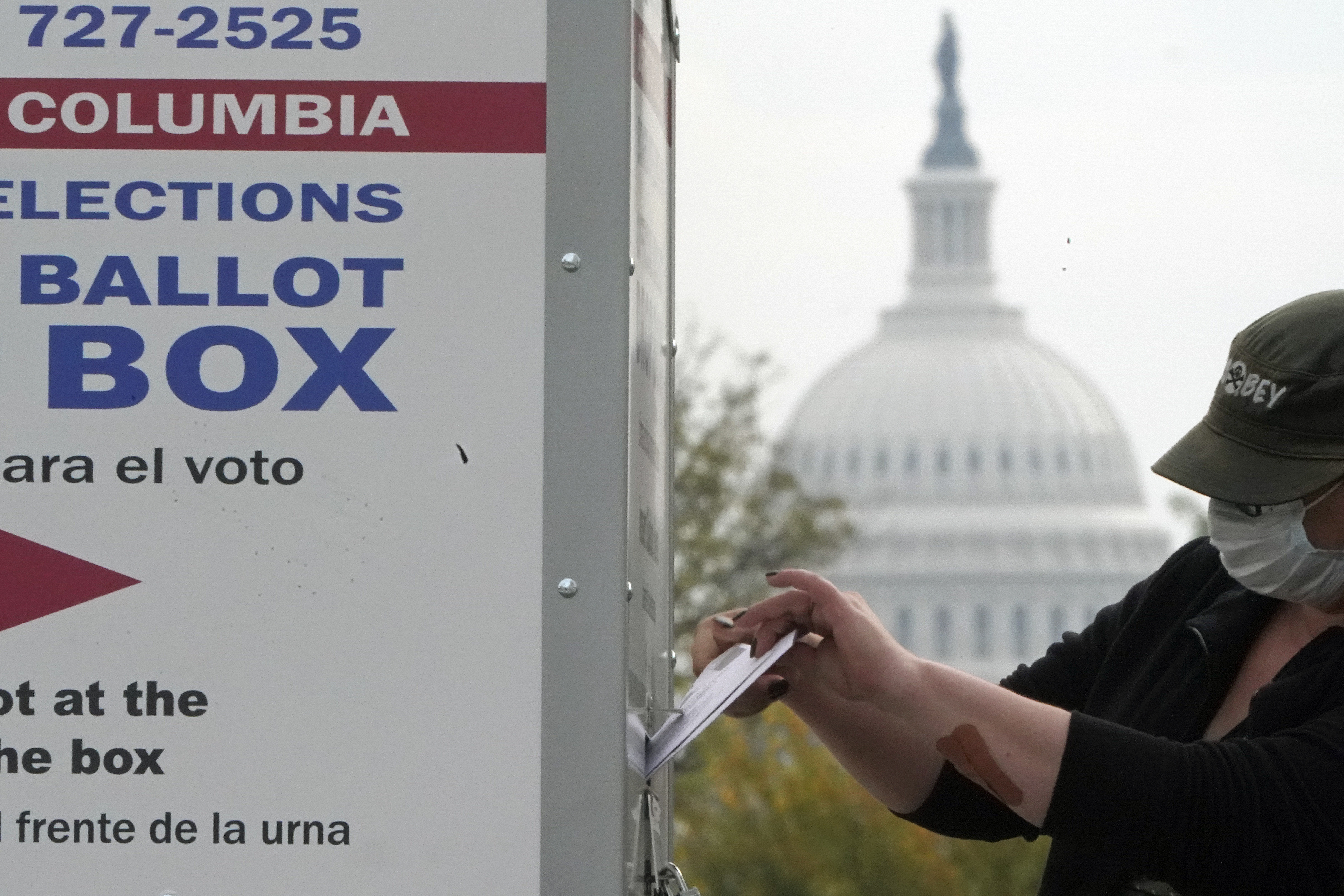 A person in a baseball cap slides a ballot into an outdoor ballot box, with the US Capitol in the background.