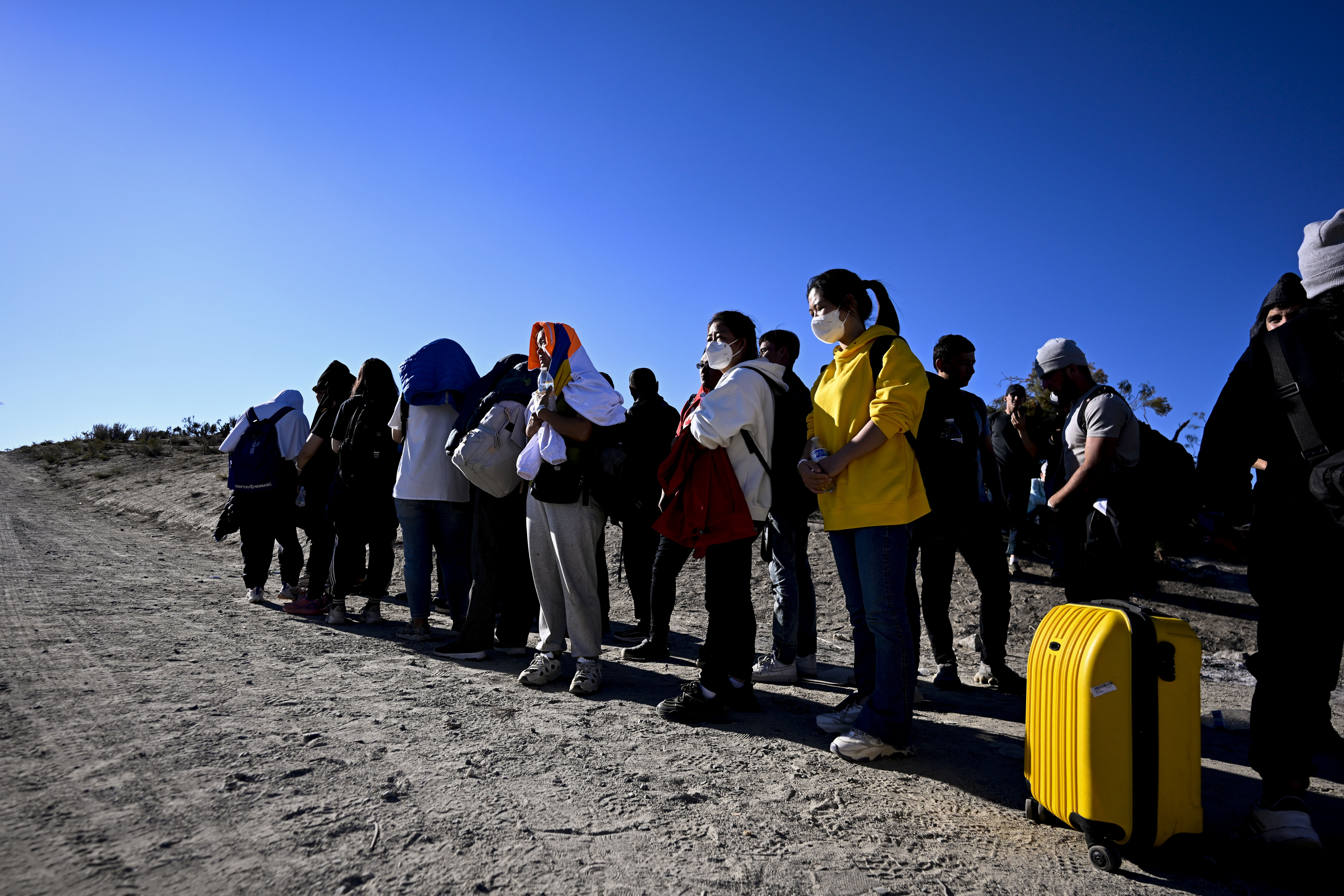 Asylum-seekers from China wait to be processed by U.S. Border Patrol agents after crossing the nearby border with Mexico. They are standing in a line, Some have luggage. One has a bright yellow suitcase