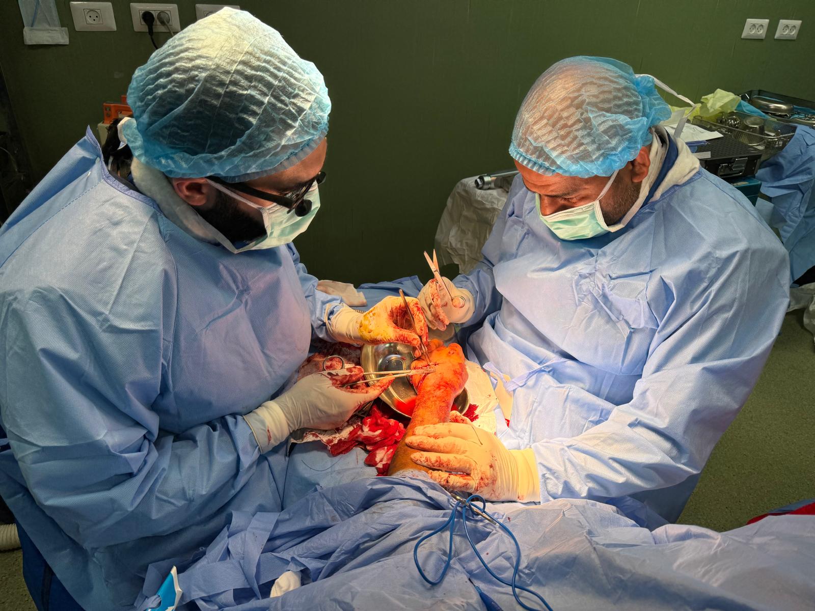 Omar Sabha and Jawad Khan, dressed in blue medical scrubs and hair nets, operate on a patient's hand, stitching it up. Blood is visible on the medical worker's gloves.