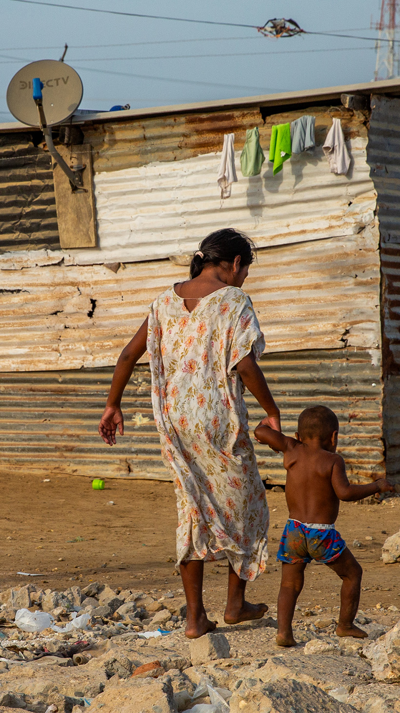 A woman and child walk hand in hand through the drylands of La Guajira, a ramshackle house in the background with laundry hanging under its roof.