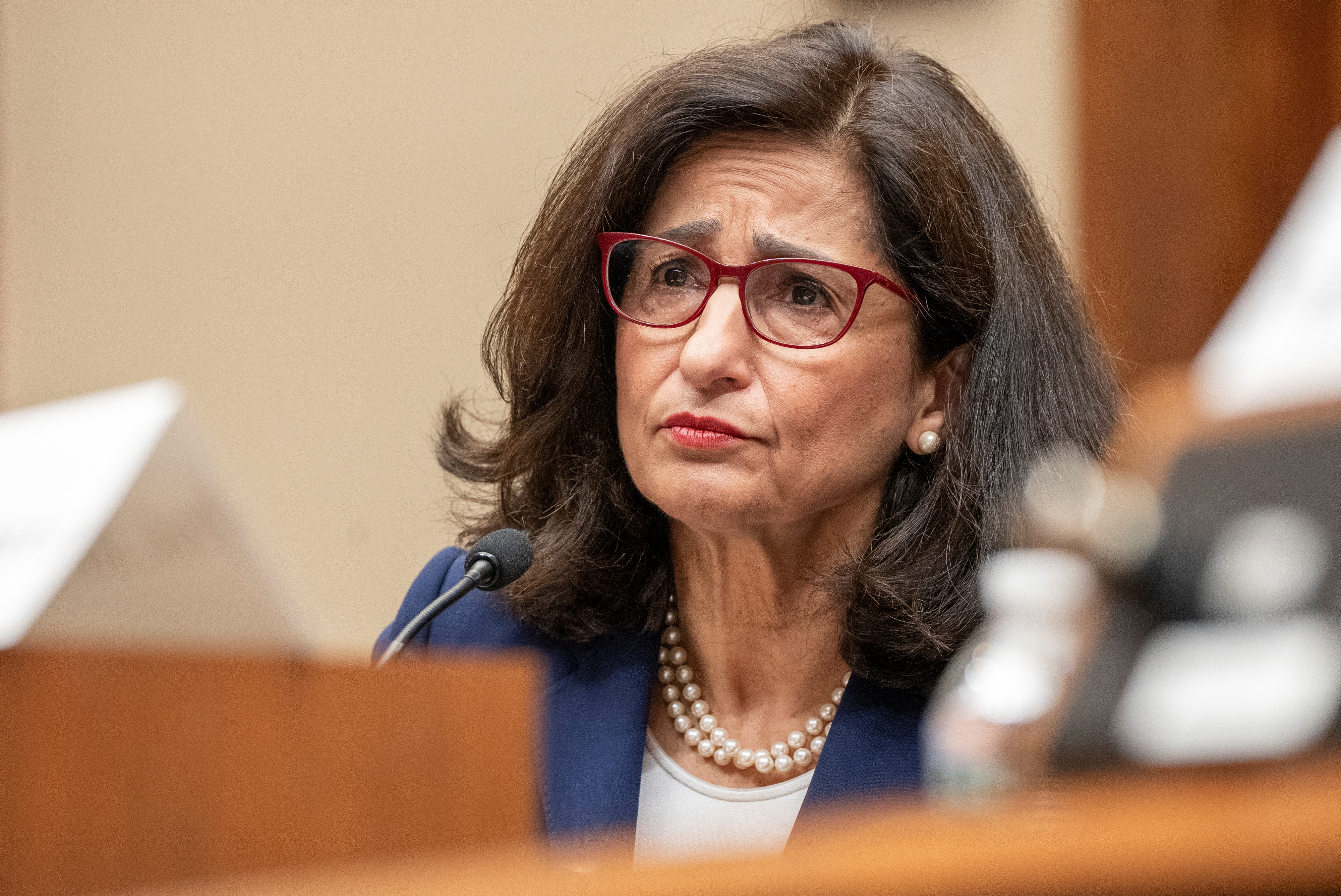 Minouche Shafik. She has dark mid-length hair and is wearing red glasses.