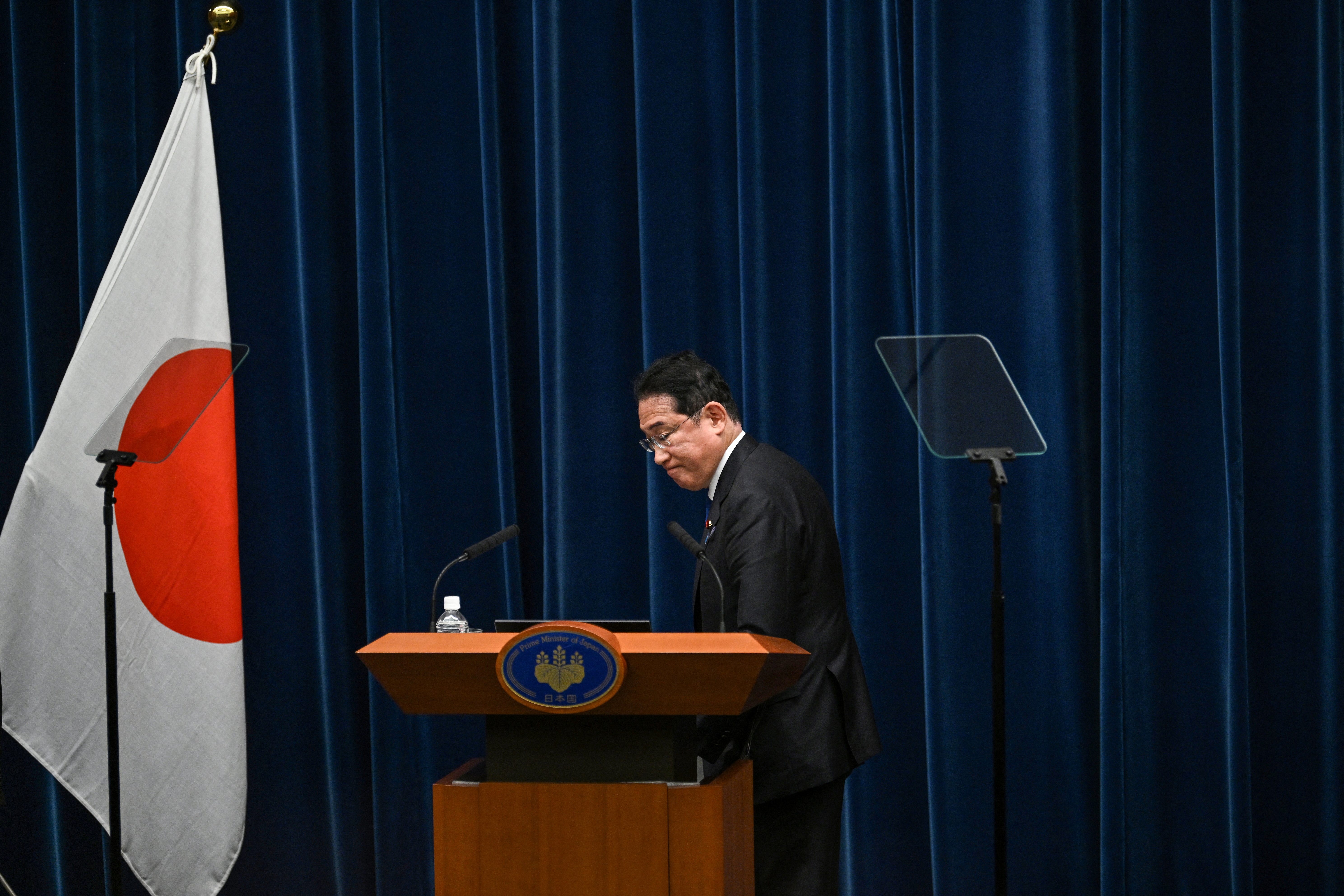 Fumio Kishida preparing to leave the stage after a press conference. He has his head bowed and is in front of a dark blue curtain. There's a Japanese flag to one side.