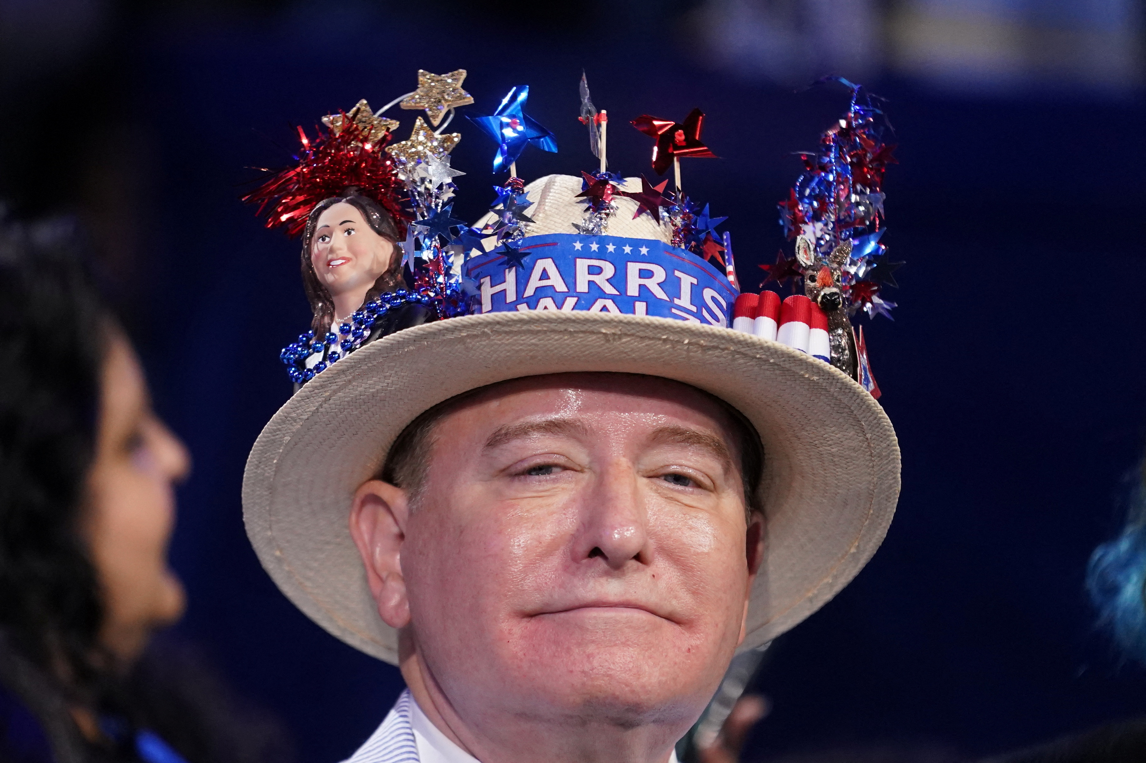 A man wears a hat decorated with sparkly stars and a sash that says, "Harris Walz"