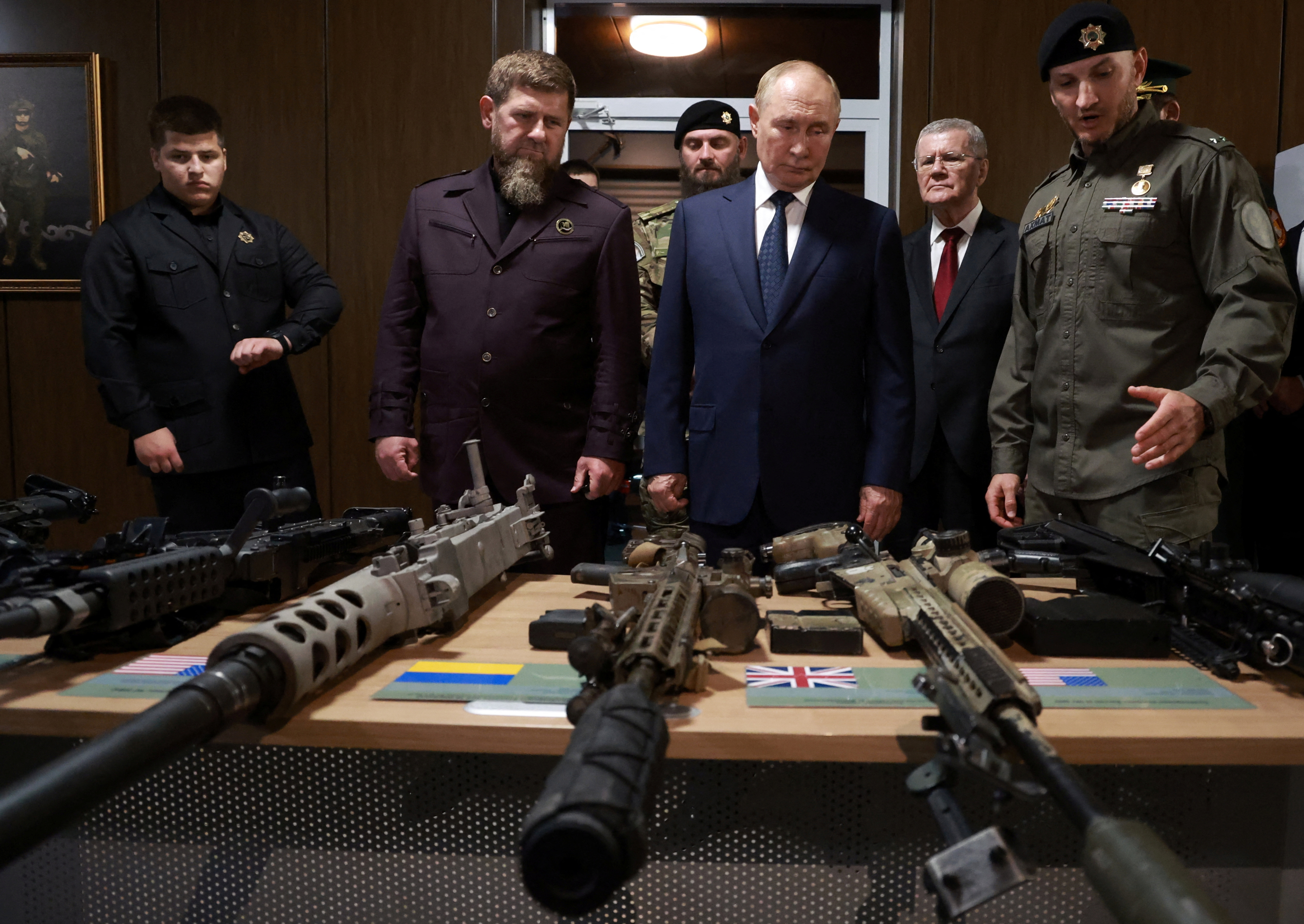Putin and Kadyrov looking at weapons. The guns are laid out on a table in front of them. Another man is speaking to Putin