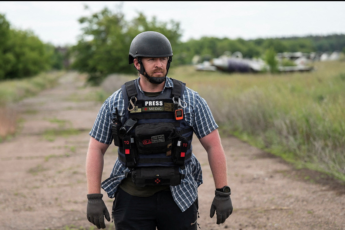 Reuters safety adviser Ryan Evans. He is standing in a field in Ukraine. He is wearing body armour and a hard hat. HIs vest has patches reading press and medic.