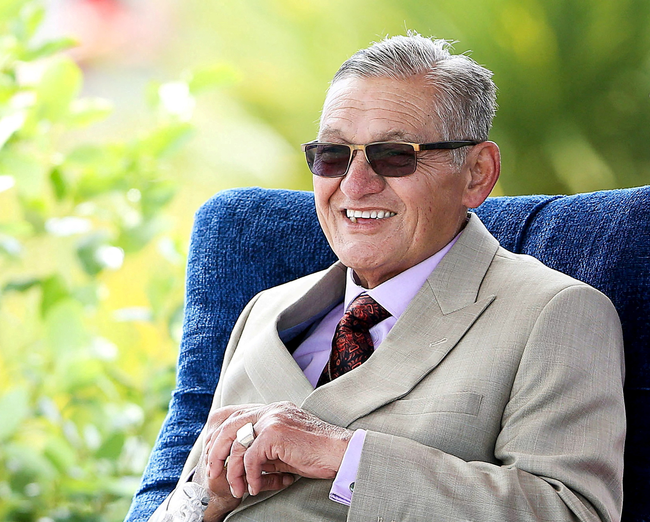 Maori King Tuheitia. He has grey hair and is wearing a light beige suit. He is also wearing sunglasses. He is sitting in a chair. He is smiling.