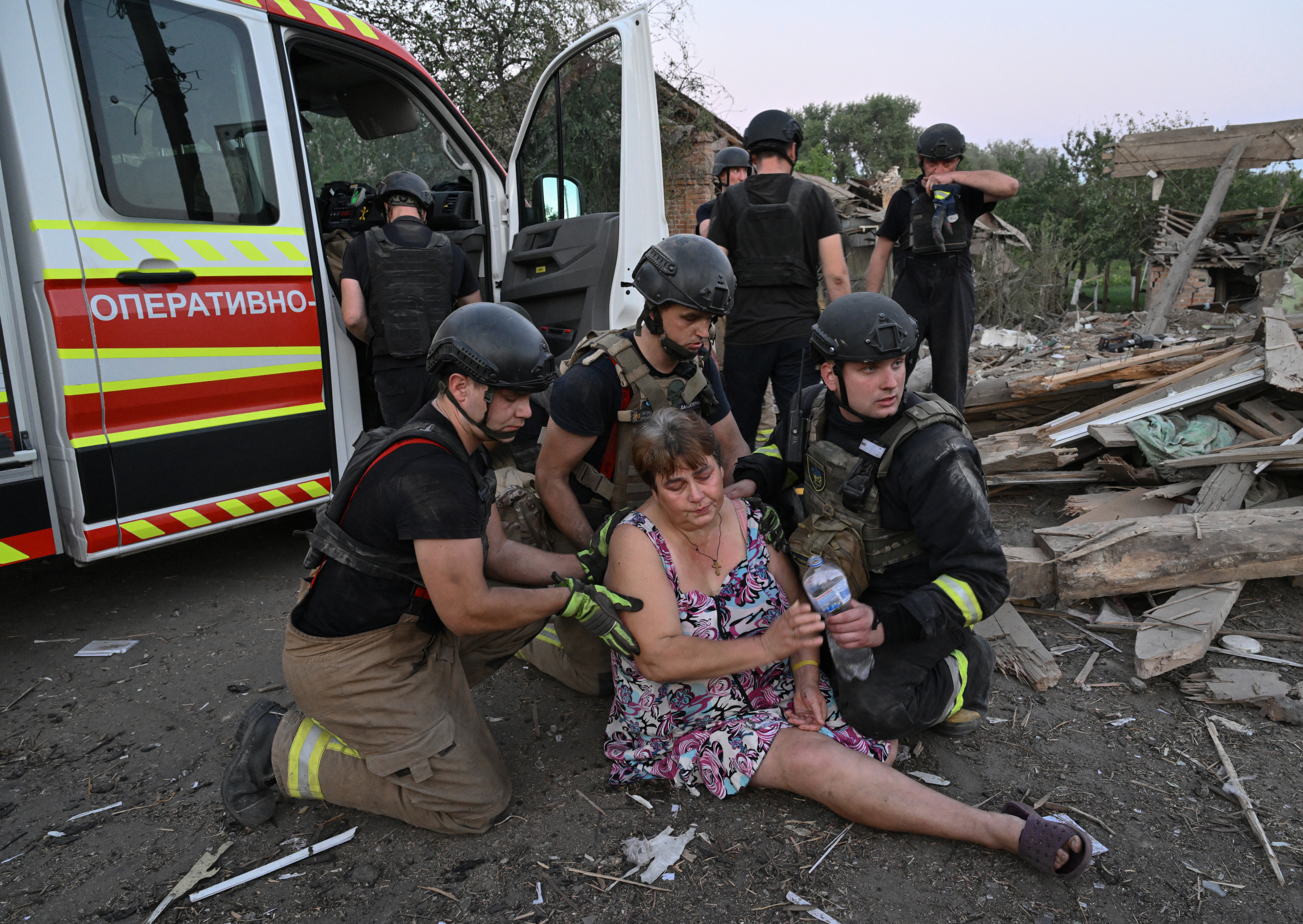 A Ukrainian woman being helped by resue teams after she fainted. An ambulance is behind her. There is lots of rubble around.