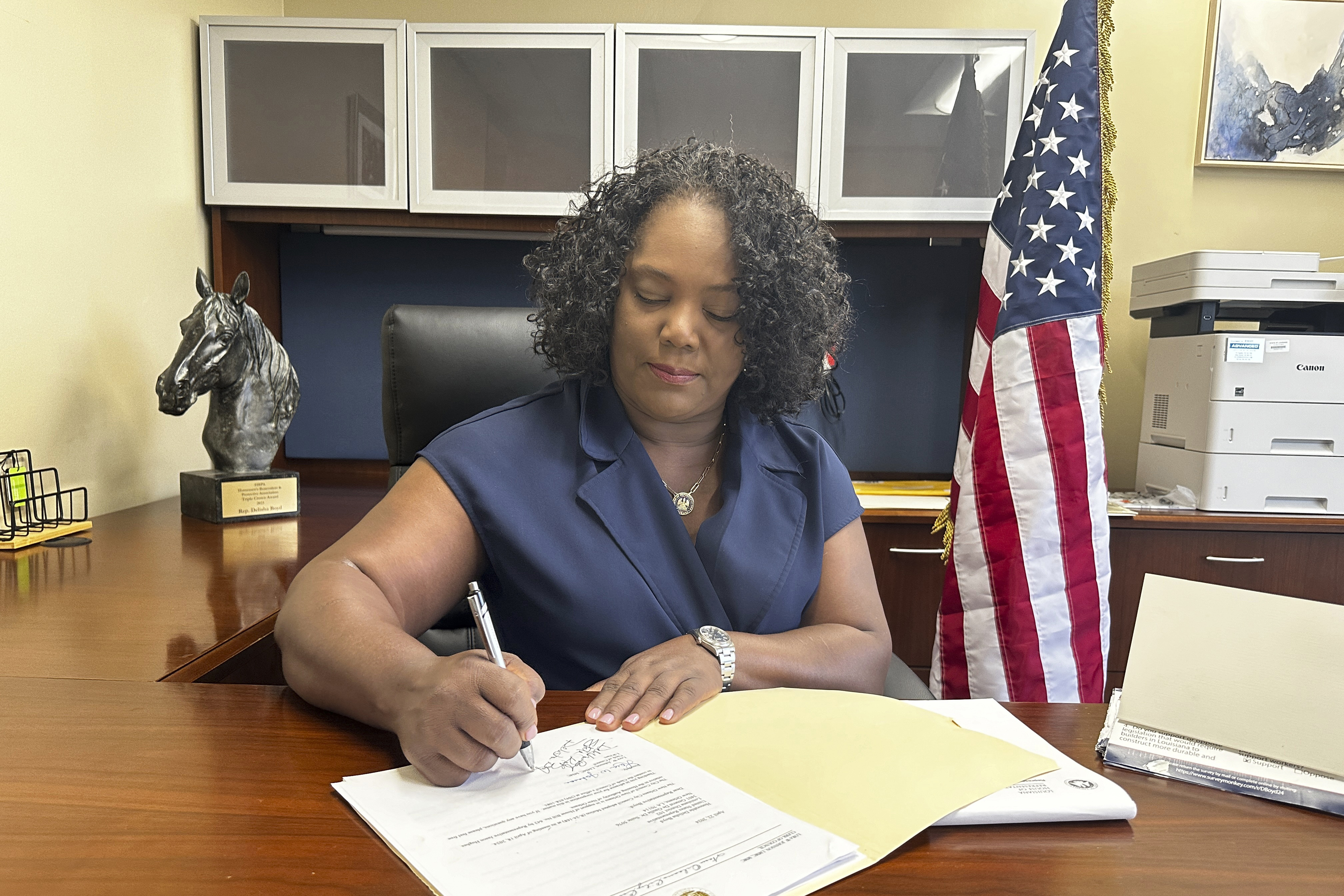 Delisha Boyd signs a piece of paper in a folder in her office. Behind her desk sits a US flag.