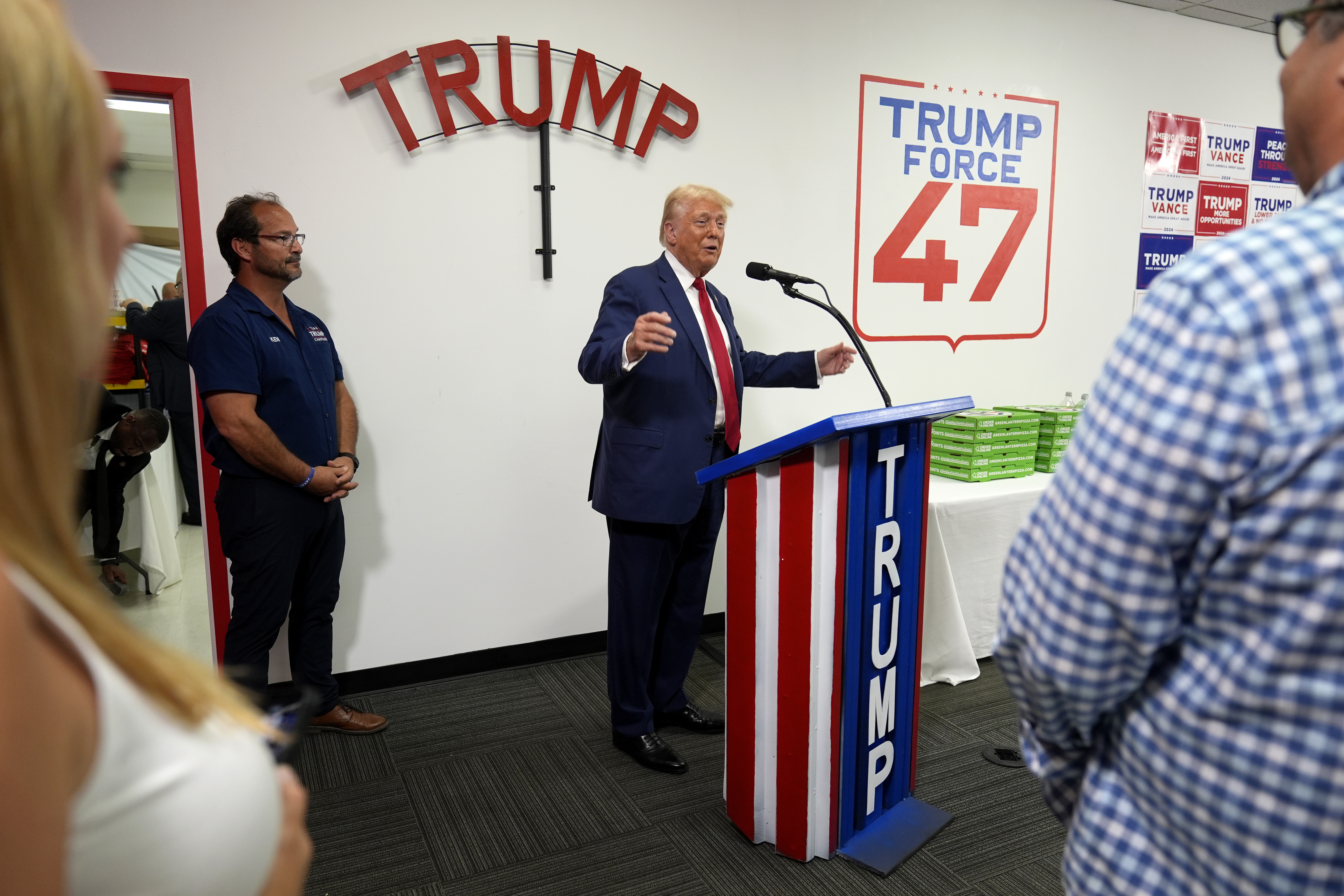 Donald Trump campaigns at a US flag-themed podium, under a sign with his name.