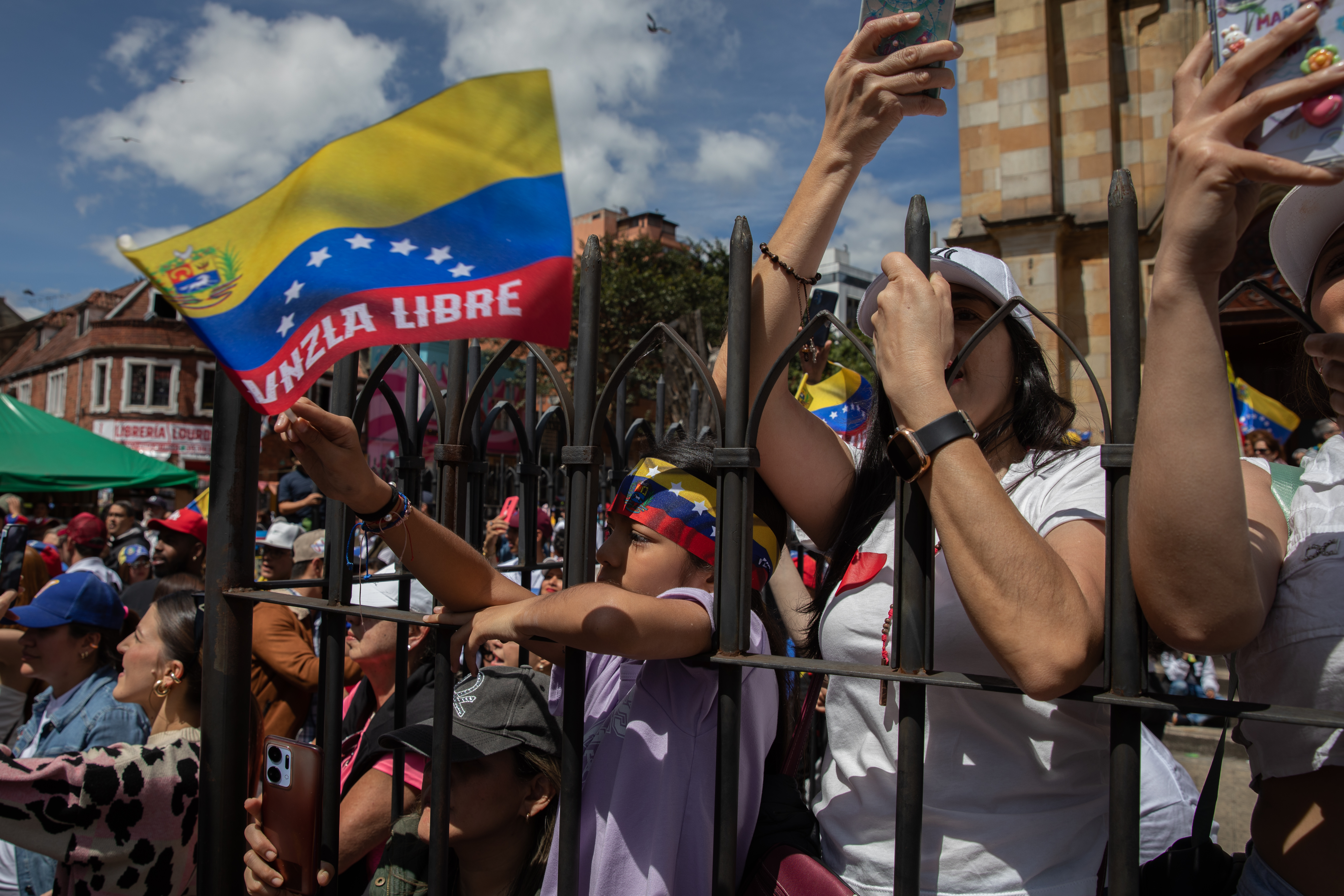 Protesters in Bogota press themselves against a metal fence, waving Venezuelan flags and holding up their cellphones to record.