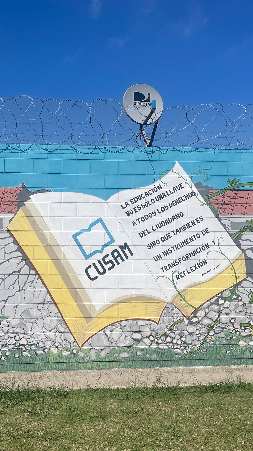 A mural on a cinderblock wall — below satellite dishes and barbed wire — shows an open book with the word "CUSAM" inside as well as a phrase: "La educacion no es solo una llave a todos los derechos del cuidadano, sino que tambien es un instrumento de transformacion y reflexion.'