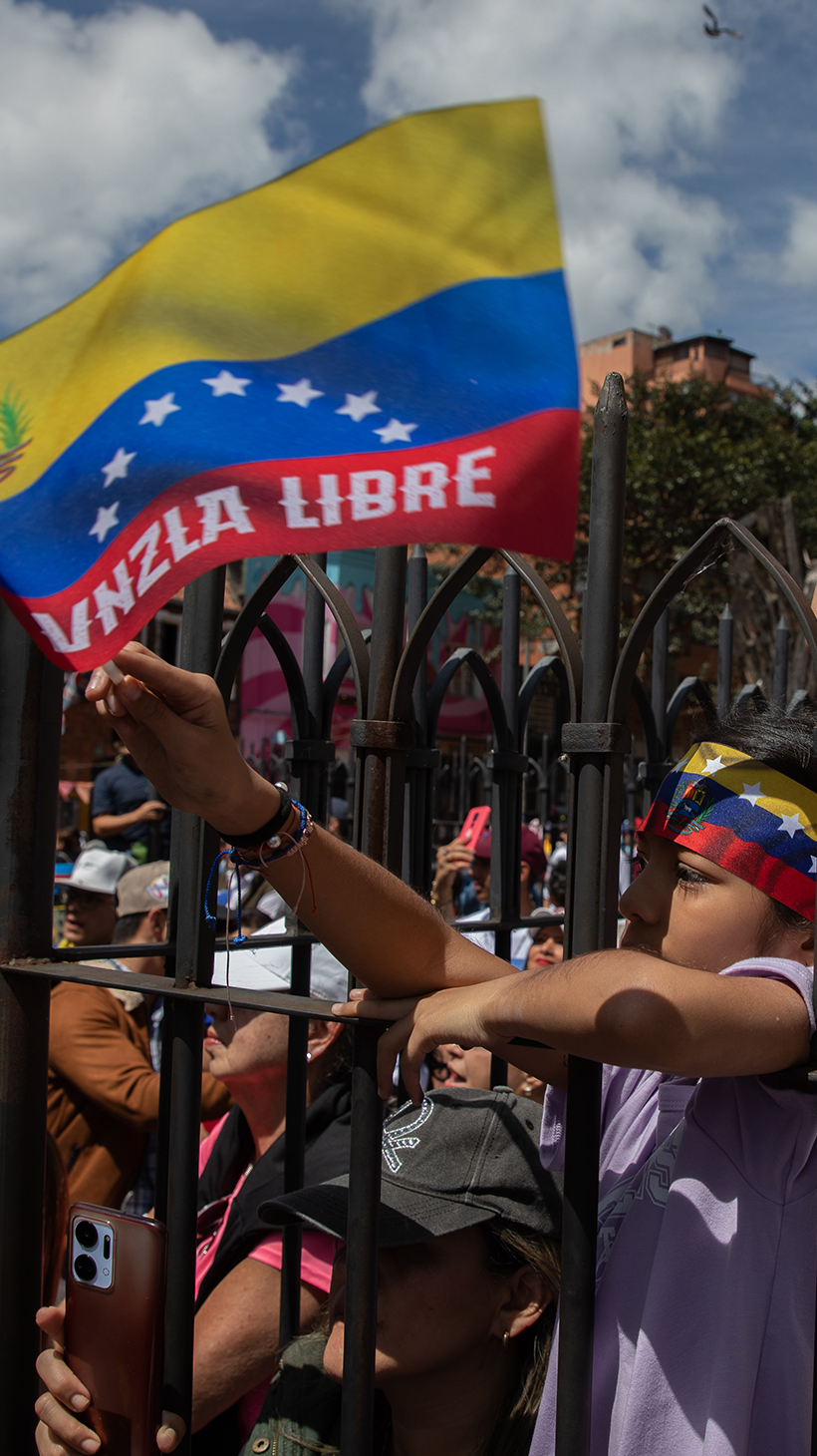 Protesters in Bogota press themselves against a metal fence, waving Venezuelan flags and holding up their cellphones to record.