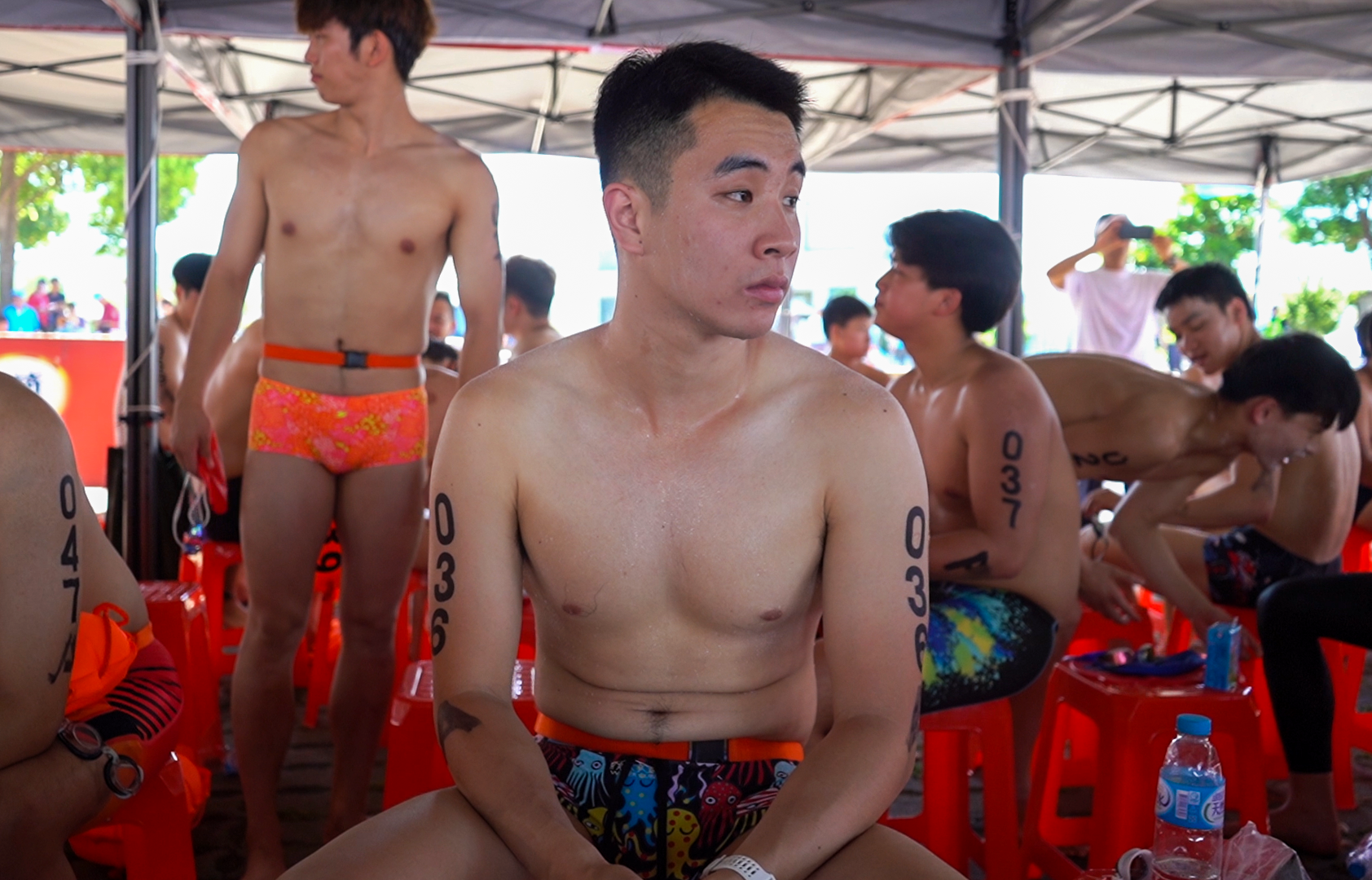 Swimmer Liu Xi Jiu. He is in his swimming shorts and seated. He looks pensive. Other swimmers are behind him