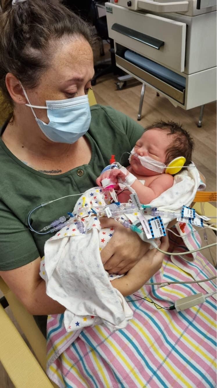 Sam Hall cradling her newborn son in hospital. She is wearing a face mask. He has lots of tubes attached to his little body