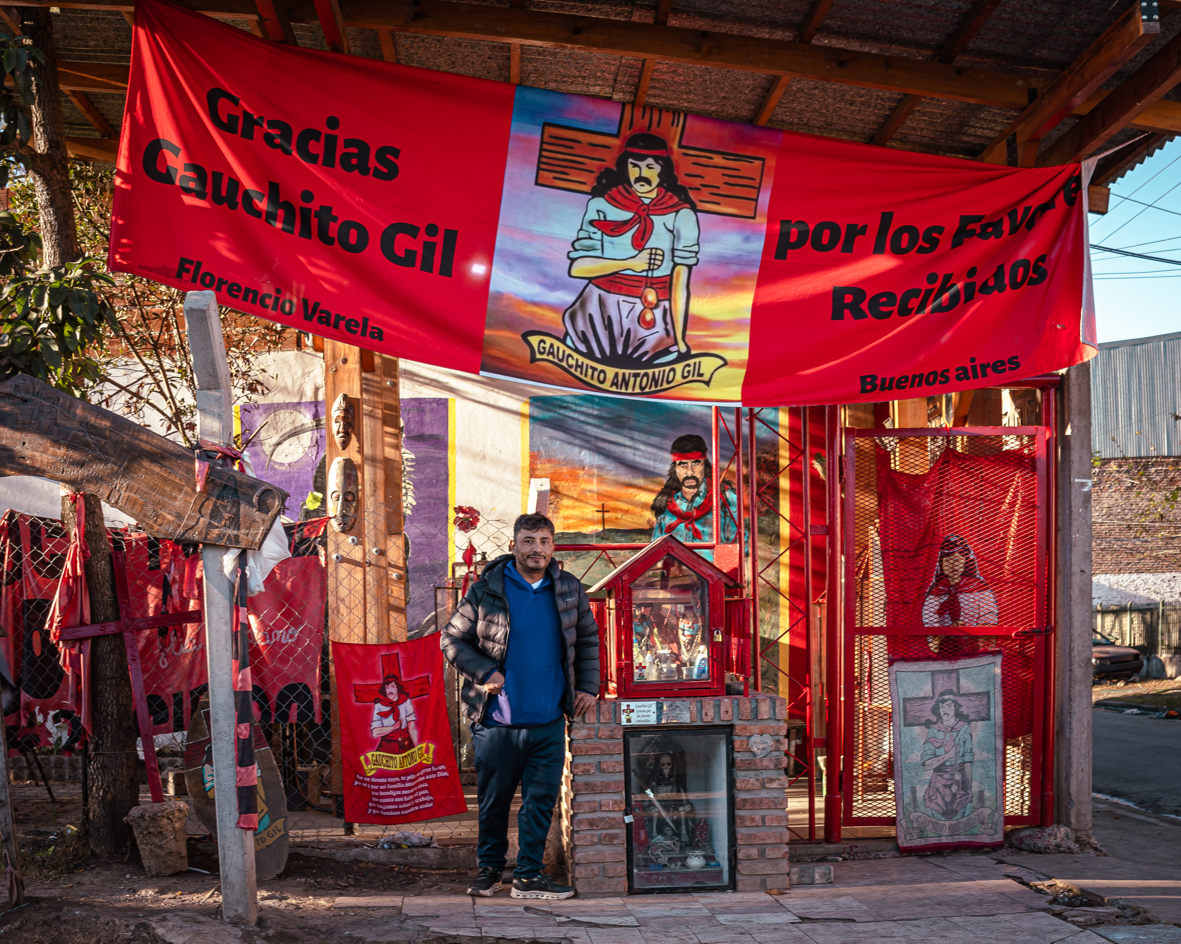 Waldemar Cubilla poses next to a small shrine, situated beneath a large red banner celebrating a local folk hero, Gauchito Gil.