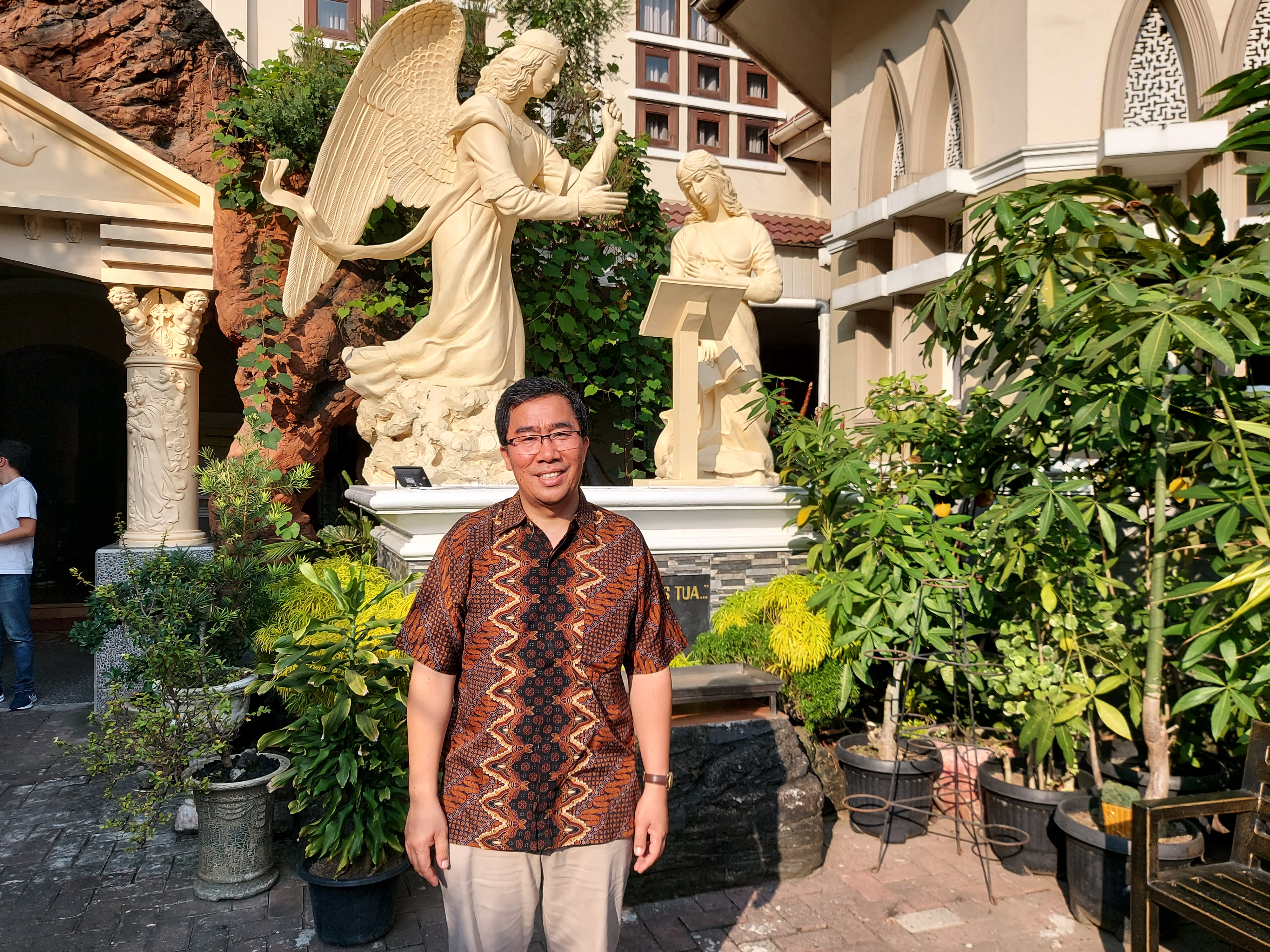 Father Joseph Gultom standing outside the church. He is wearing a brown batik shirt. He's smiling.