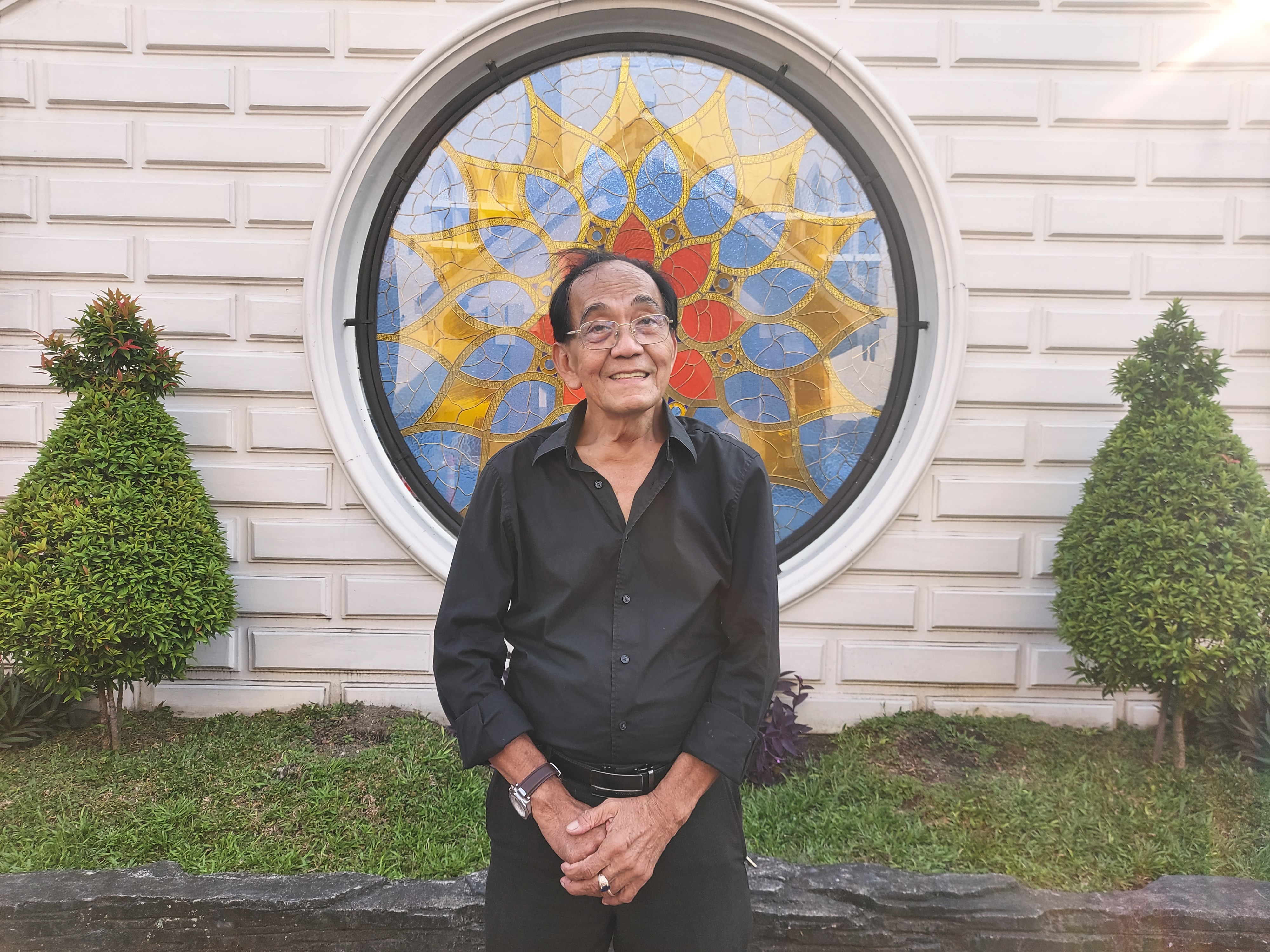 Erwin, an Indonesian Catholic. He is standing outside the Immaculate Conception of Mary Cathedral, He is wearing a black shirt and trousers and his hands clasped in front of him. He is smiling. 