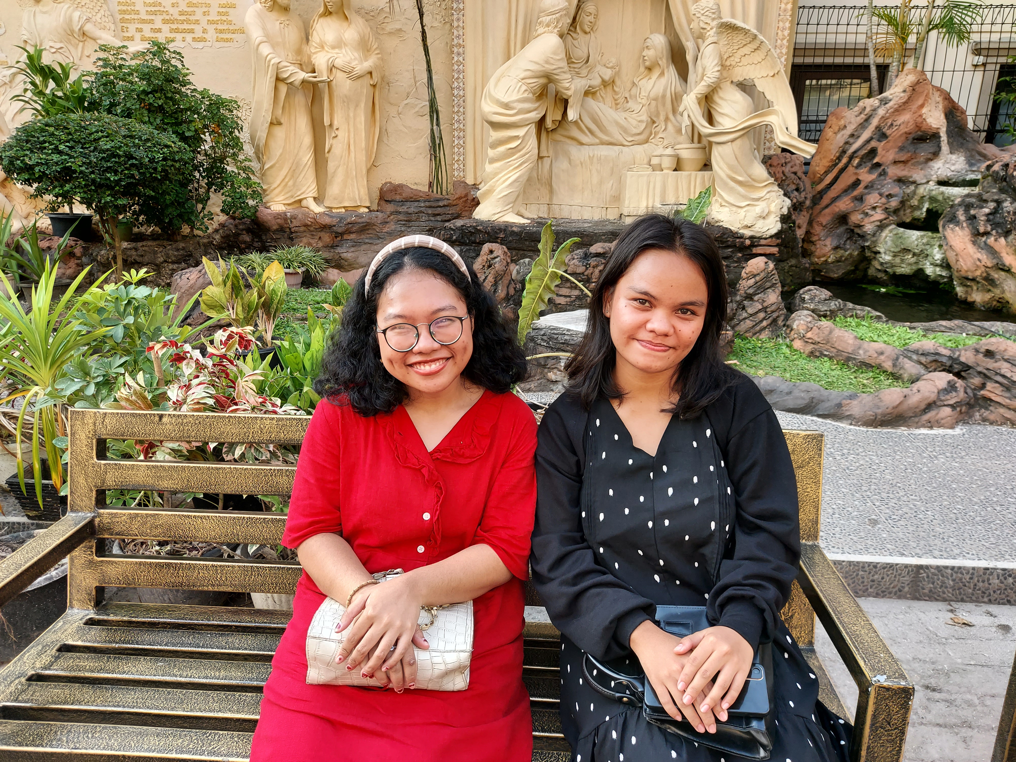 Church goers Ririn Silalhi (left) and Yola Marpaung (right) sitting on a bench outside the church. Ririn is weearinga. red skirt and has her bag on her lap with her hands folded on top. She has bobbed dark curly hair which is held back in a head band. Yola is wearing a black dress and has her hands in her lap. They are both smiling.
