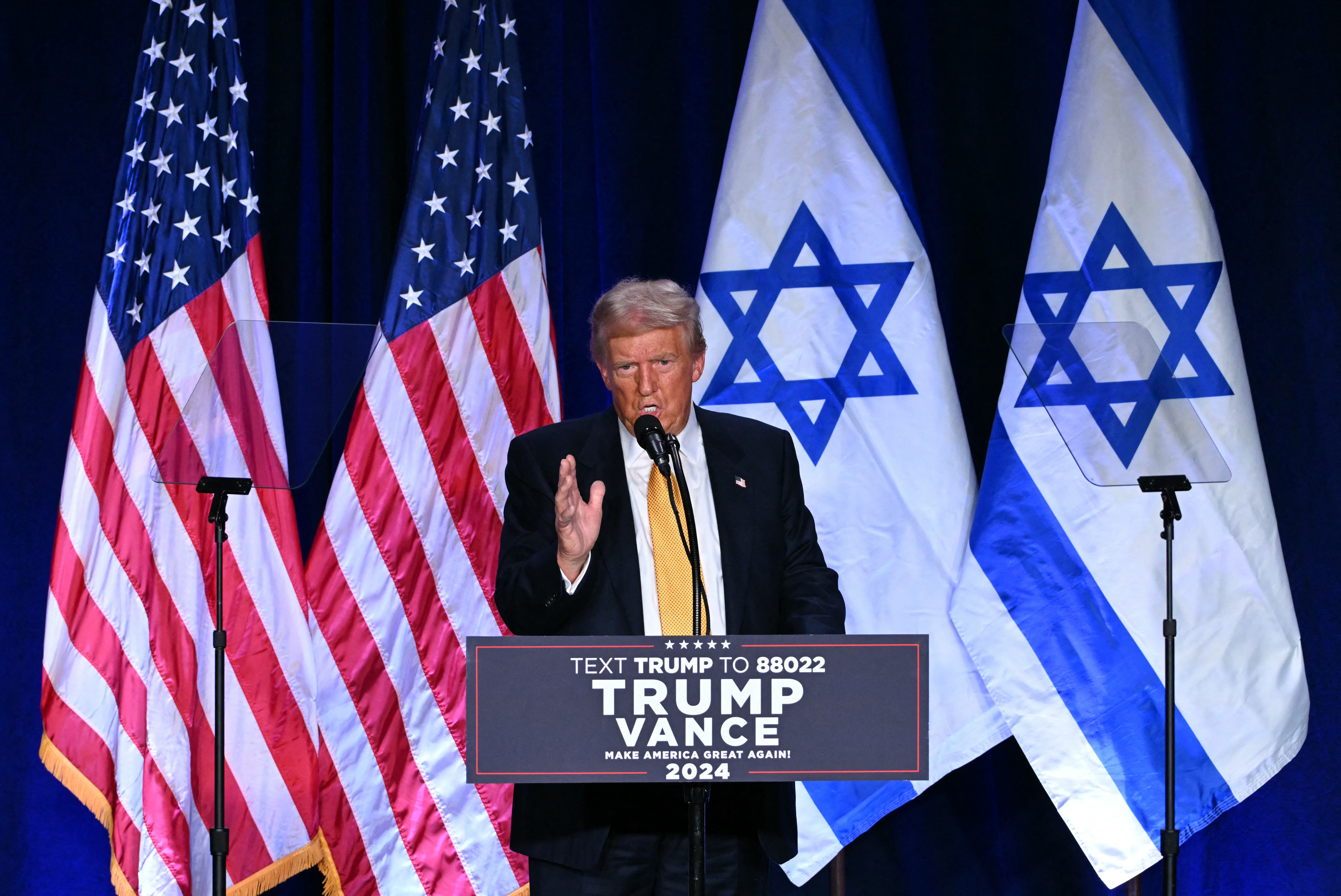 a man in a yellow tie stands in front of 2 US flags and 2 israeli flags and a podium that says trump vance