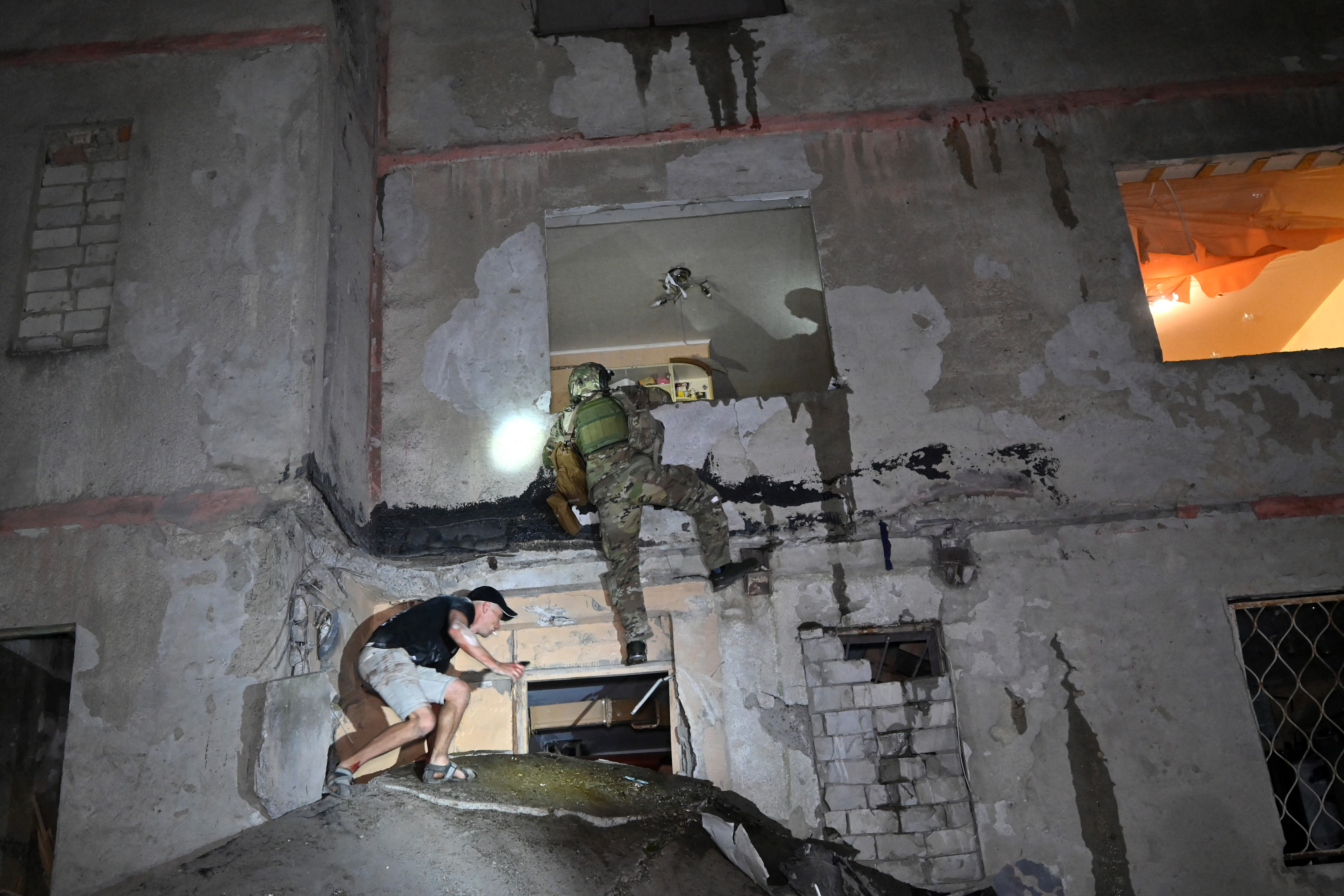 A Ukrainian law enforcement official climbing into an apartment block damaged in a Russian attack. A local resident is clambering over some of the rubble beneath to get into the building. One of the rooms is on fire,