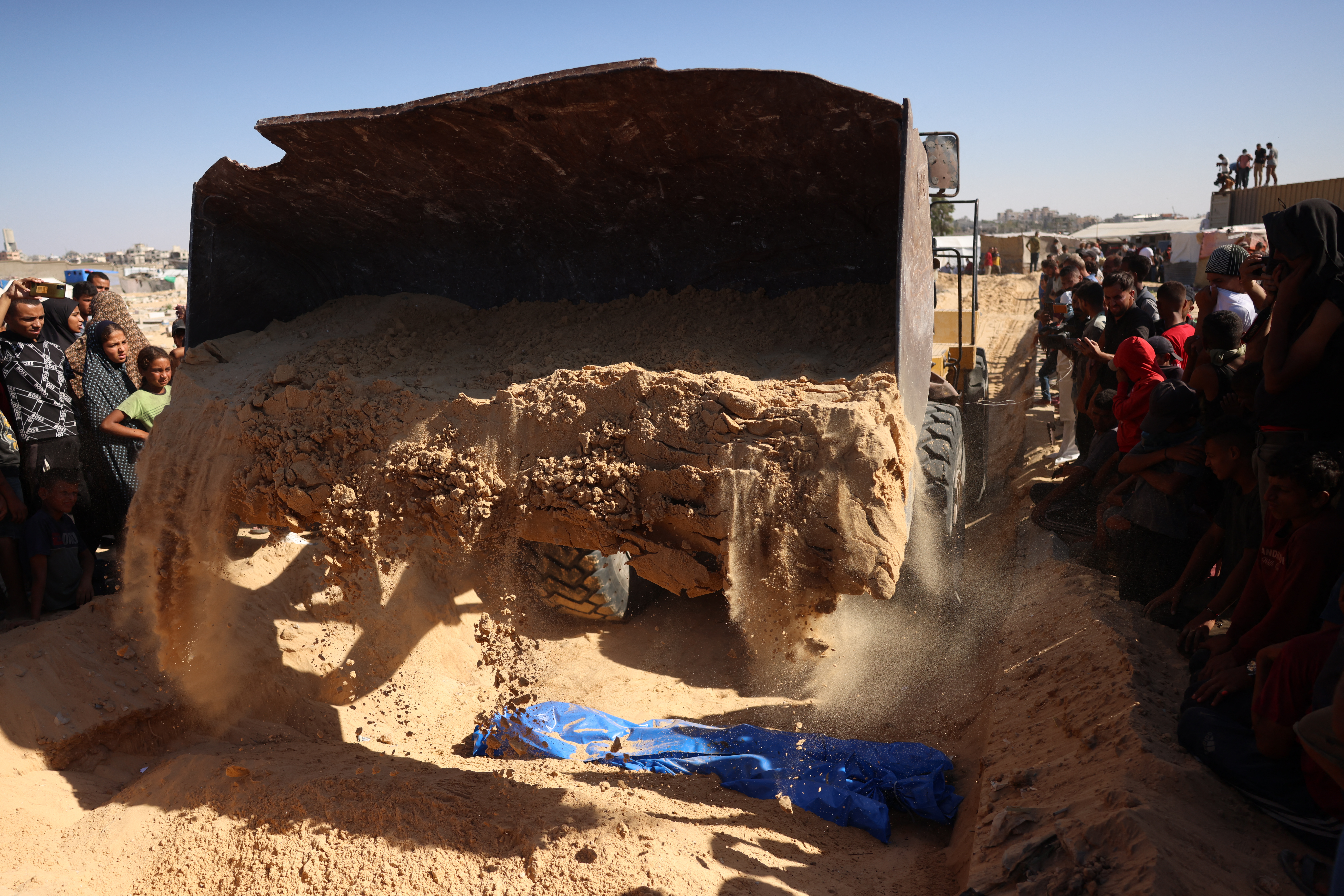 People watch as an excavator pours sand over one of the 88 bodies buried in a mass grave in Khan Yunis